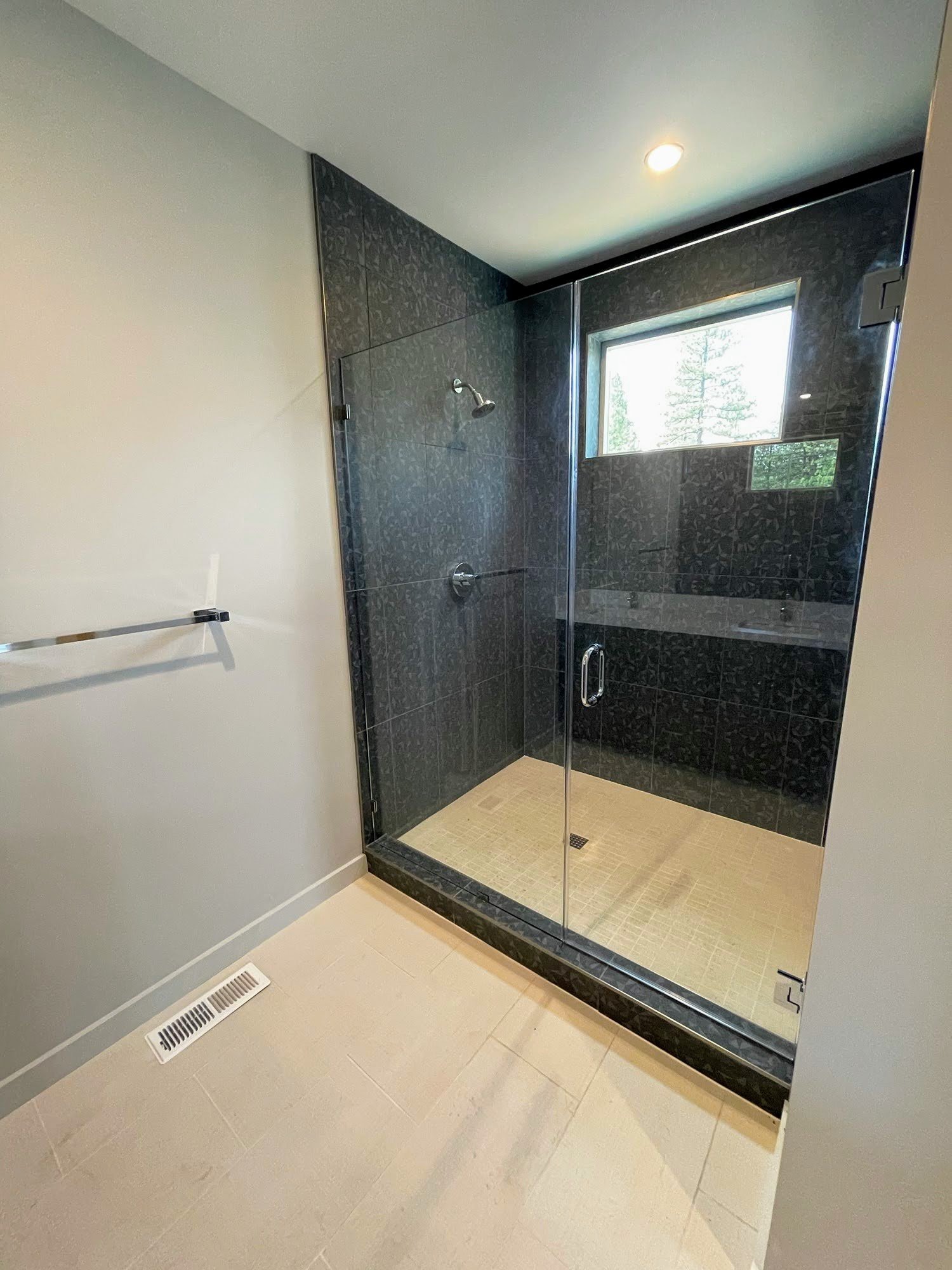 Modern walk-in shower with glass enclosure, dark tiled walls, a small window, and beige tile flooring.