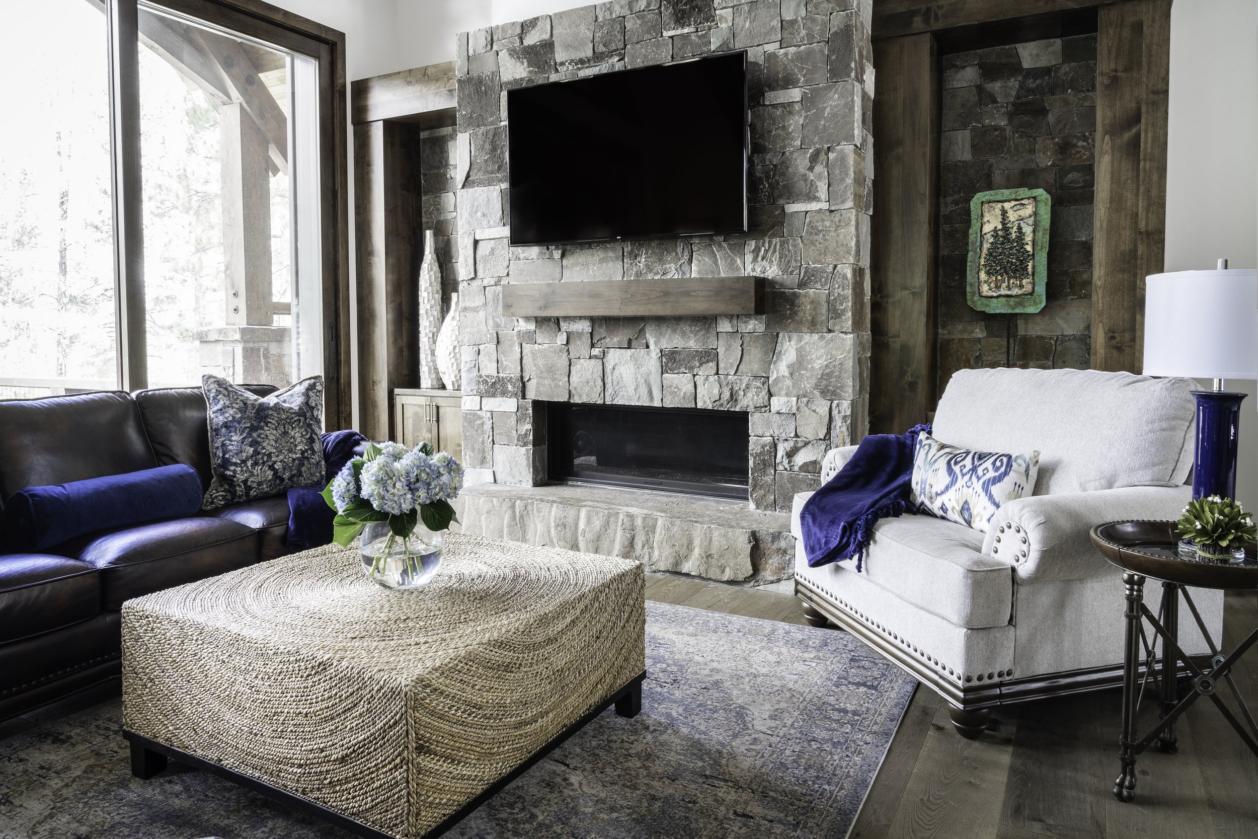 Living room with stone fireplace, wall-mounted TV above the fireplace, a cream-colored armchair with a blue blanket, a dark leather sofa with decorative pillows, a woven coffee table with a vase of blue flowers, and a side table with a plant. Large w