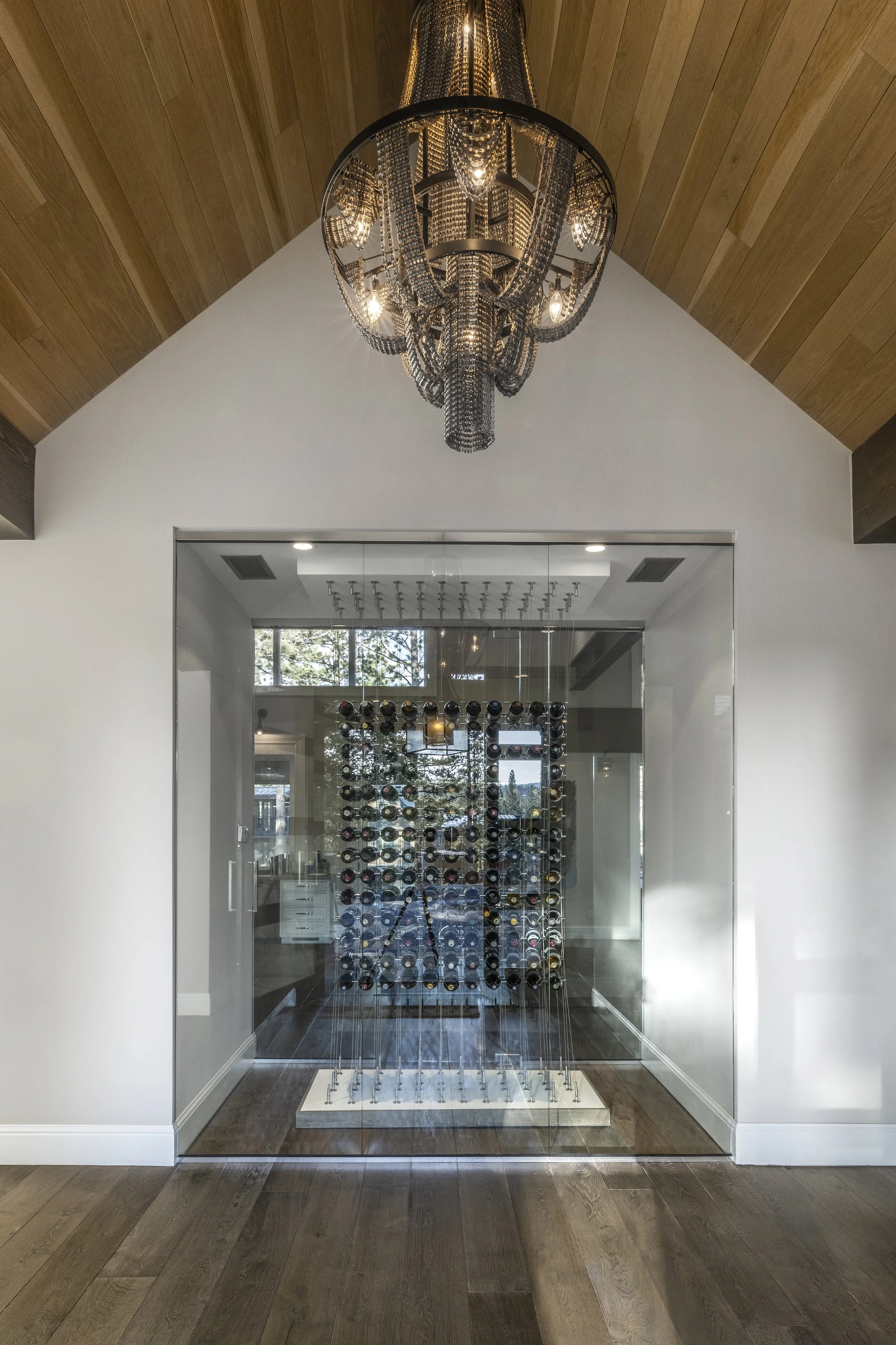 A modern wine cellar with glass walls, a white base, and a wall-mounted rack holding numerous bottles of wine. A chandelier hangs from a wooden ceiling above.