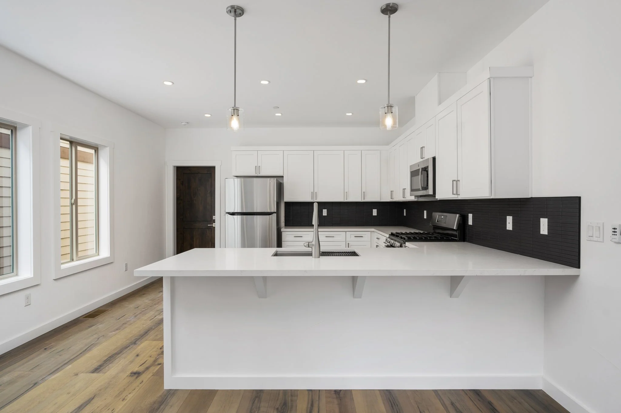 Modern kitchen with white cabinets, black backsplash, stainless steel refrigerator, oven, microwave, and island with a sink, pendant lights, and wood flooring.