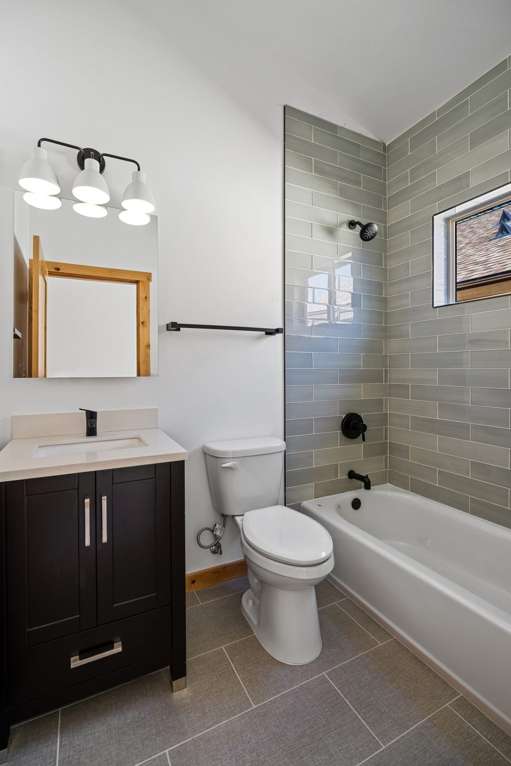Modern bathroom with a white toilet, black vanity with a sink, white mirror, four-light fixture, tan tile floor, and a shower/bathtub with gray tiled walls, a small window, and black fixtures.