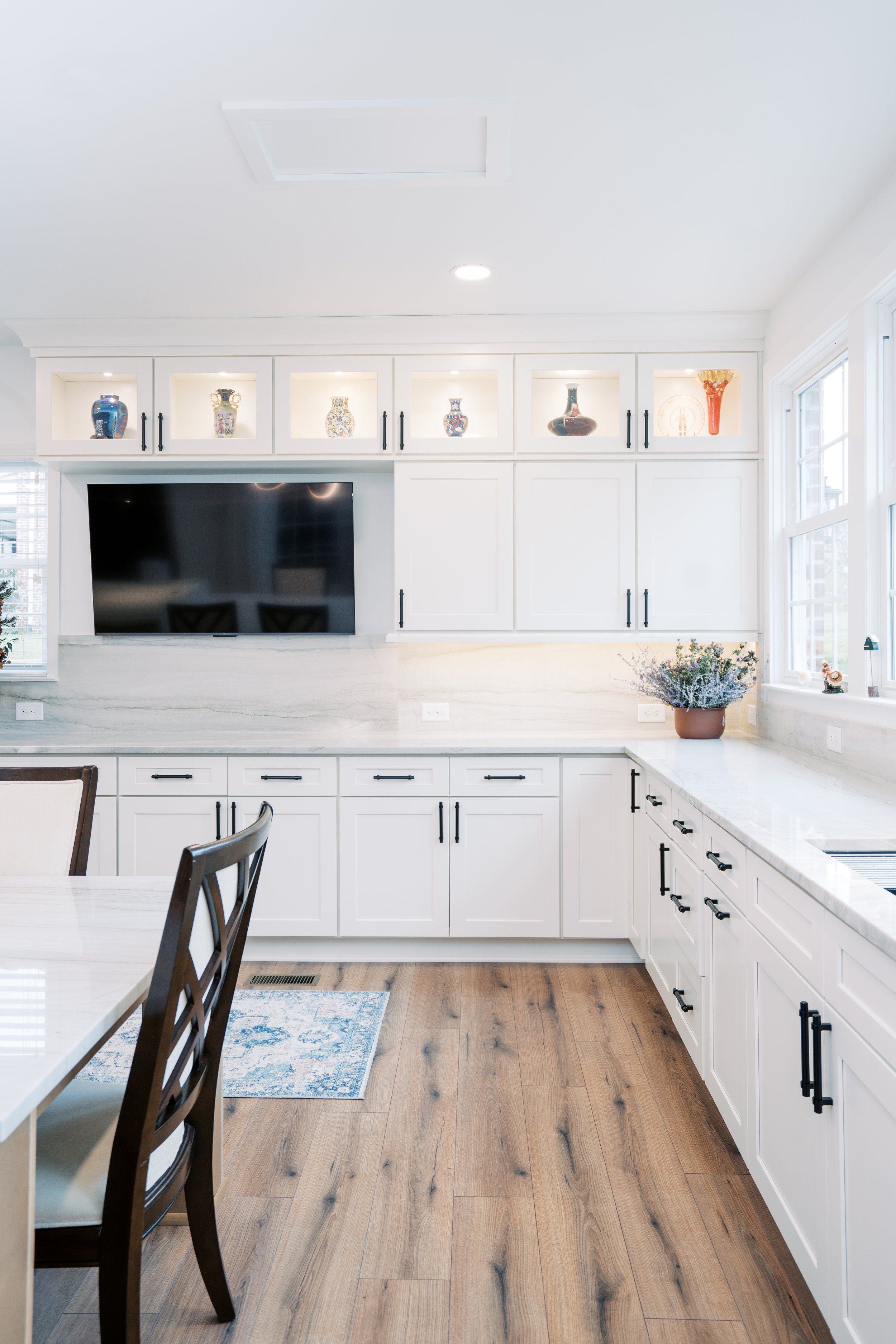 White kitchen with white cabinets, black handles, wooden flooring, a flatscreen TV on the wall, decorative vases on the upper shelf, and a window with a potted plant.
