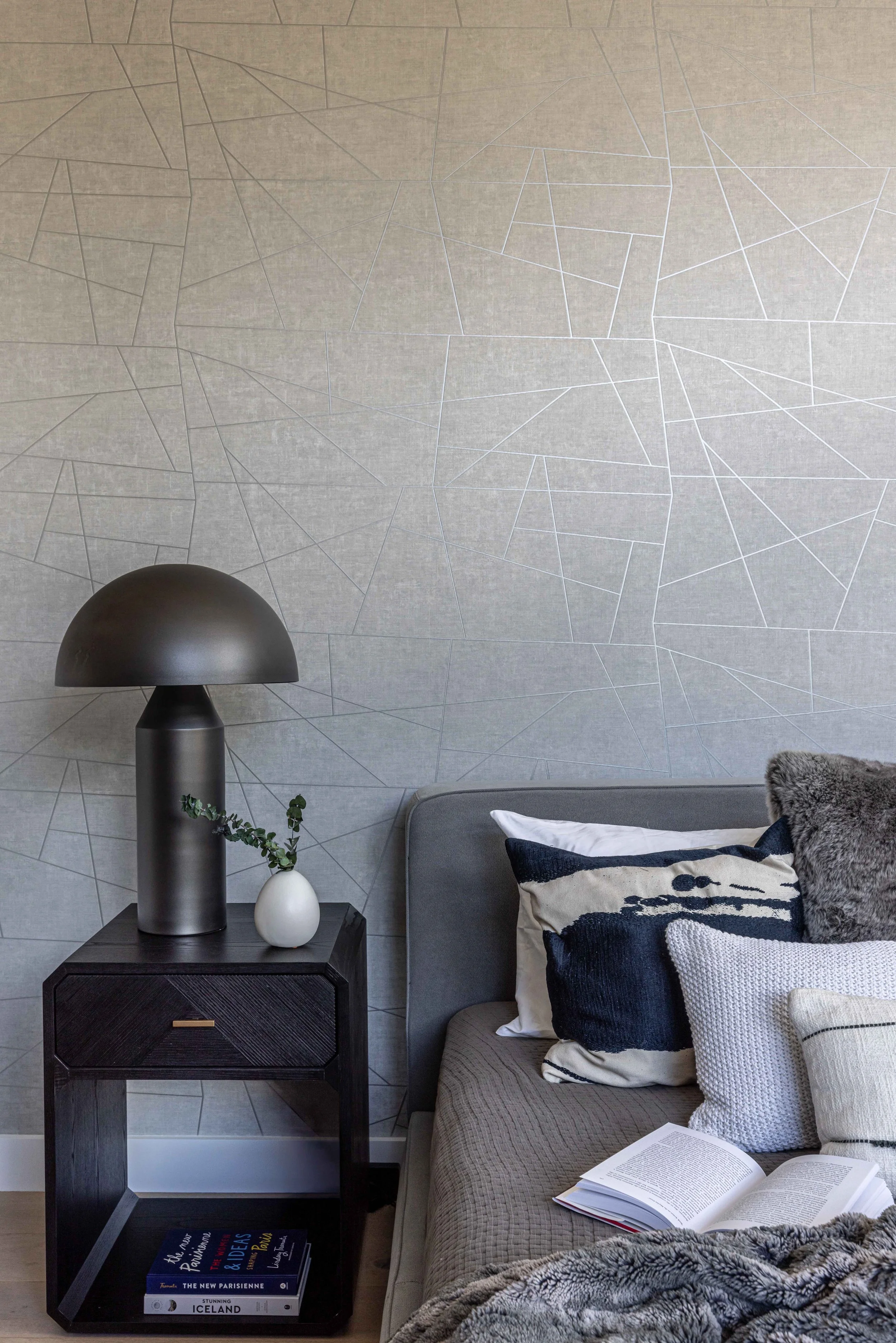 Interior of a modern living room with a grey sofa, decorative pillows, a side table with a black lamp, a small vase with greenery, and a book on the lower shelf. The wall behind features a textured geometric pattern.
