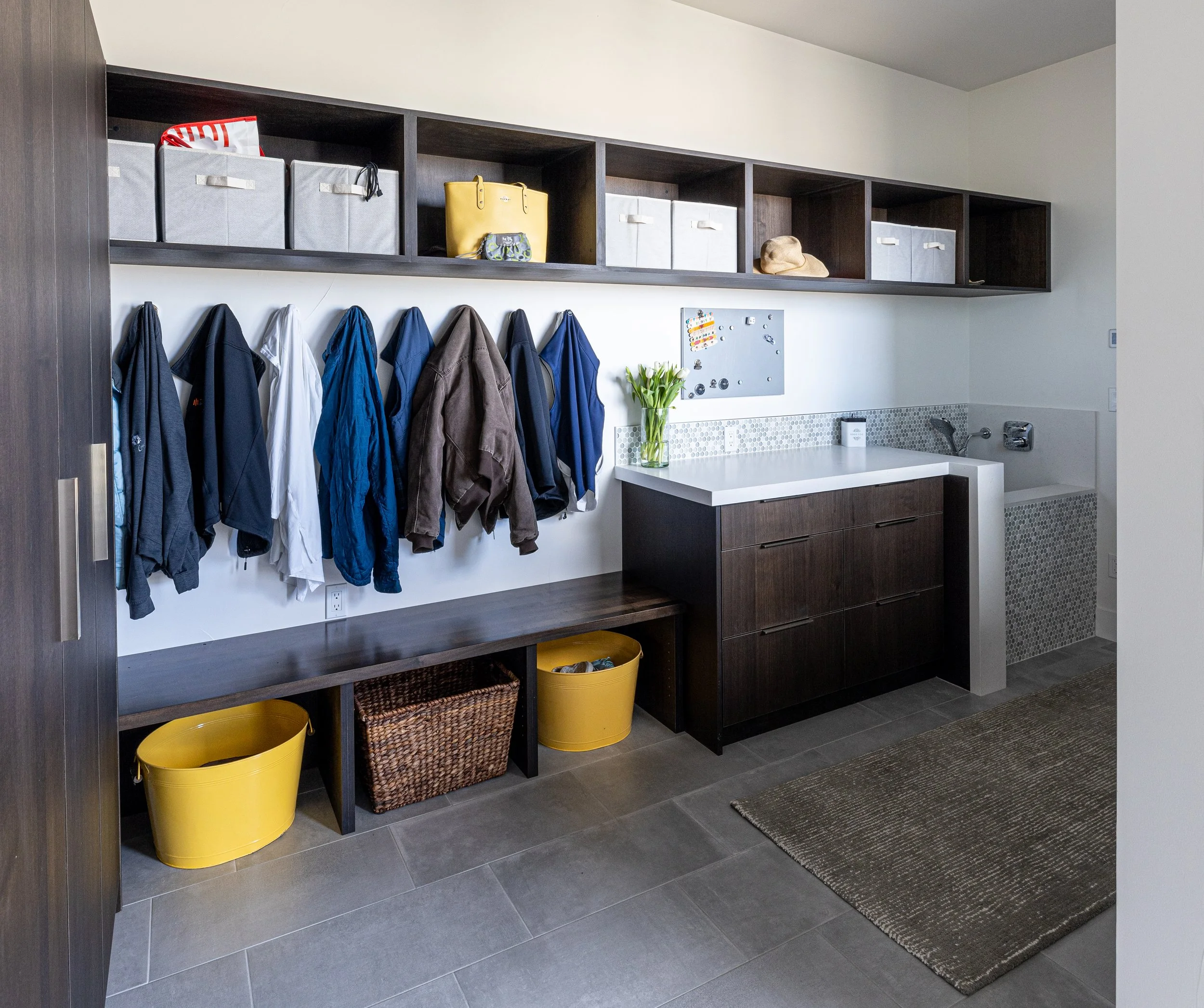 A laundry room with hanging coats on hooks, a dark wooden bench, storage bins, and a small sink area with a flower vase, with shelves holding boxes and hats.