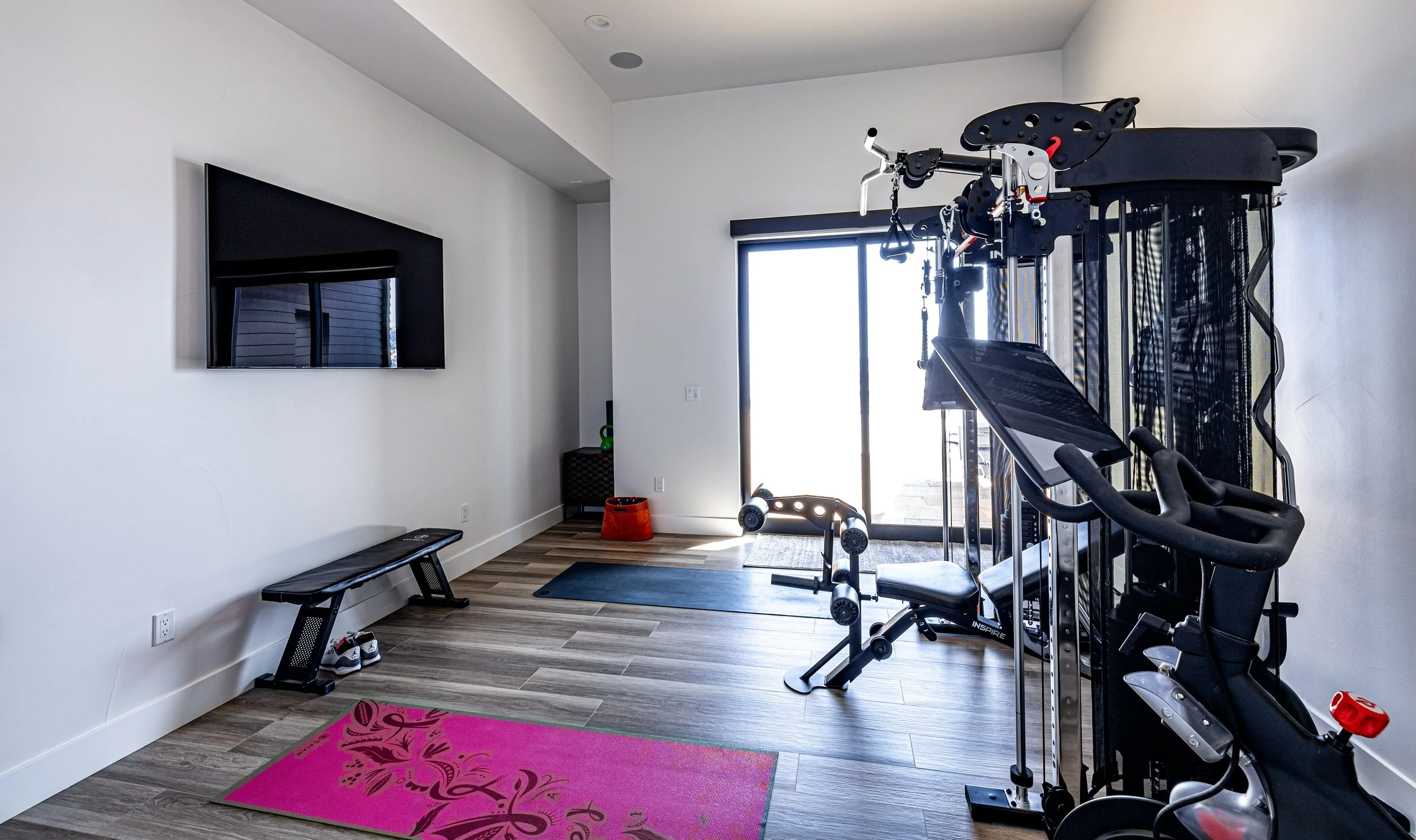 Home gym with treadmill, elliptical machine, weights, yoga mat, flat screen TV, and a sliding glass door leading outside.