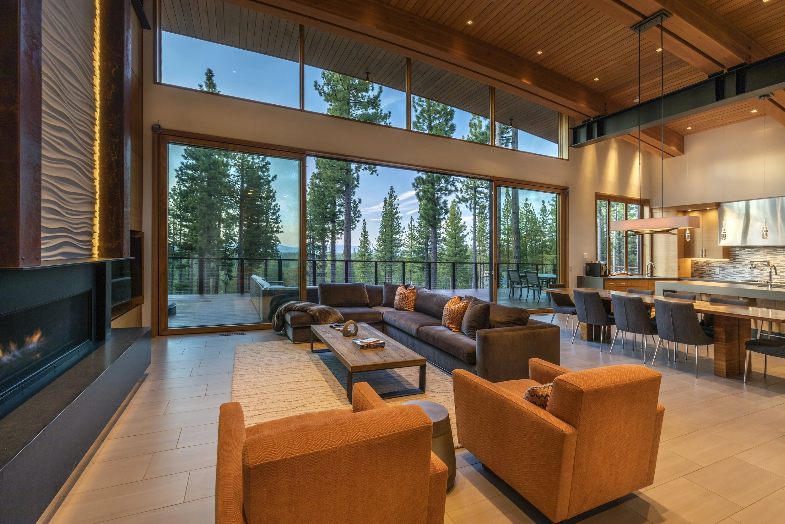 A modern living room with large glass windows overlooking tall pine trees. It has a brown sectional sofa, two orange armchairs, a wooden coffee table, a fireplace, and a kitchen/dining area with bar stools and a view of the forest outside.
