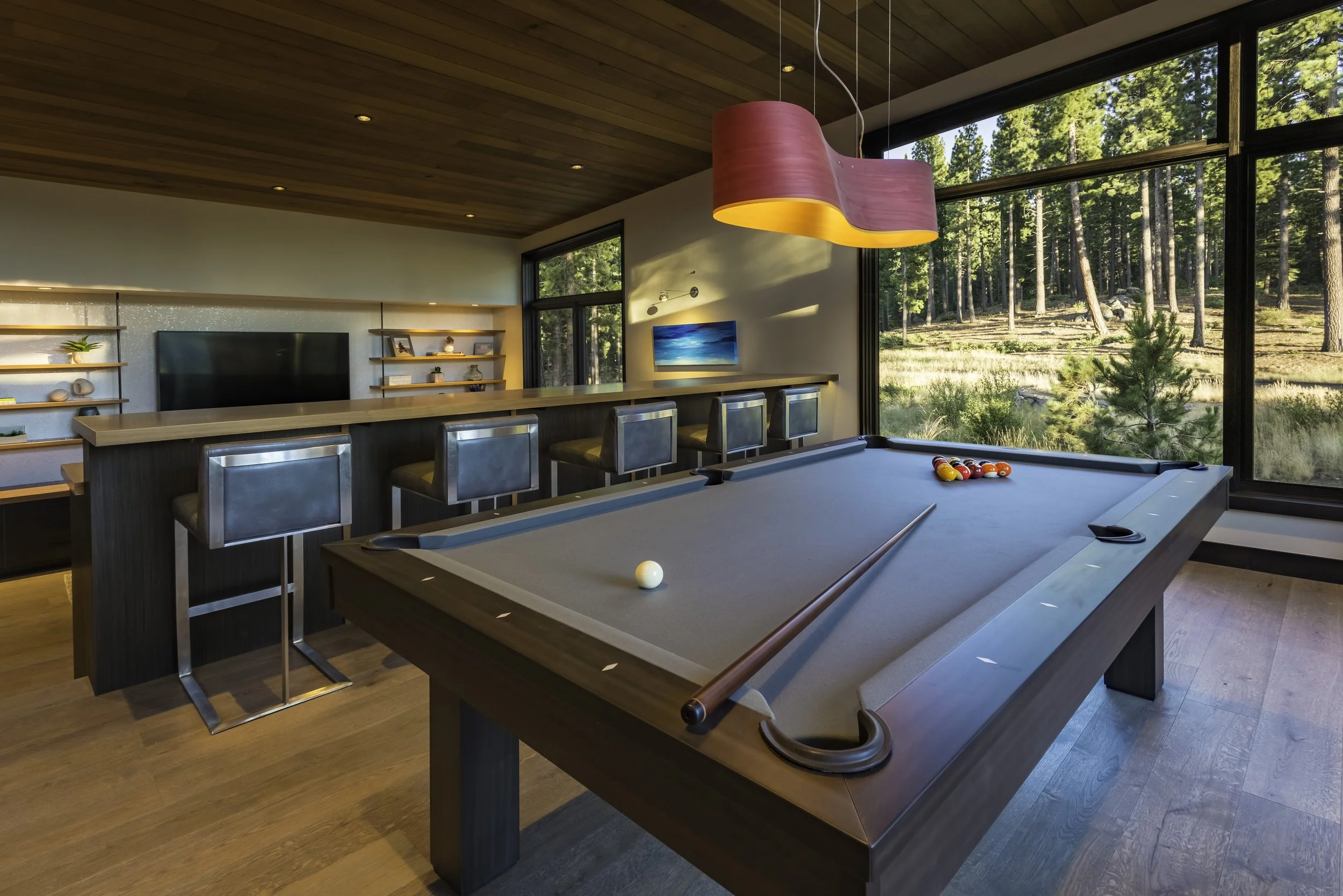 Modern recreation room with a pool table, a bar with four chairs, large windows showing a forest view, and wall shelves with decor, during daytime.
