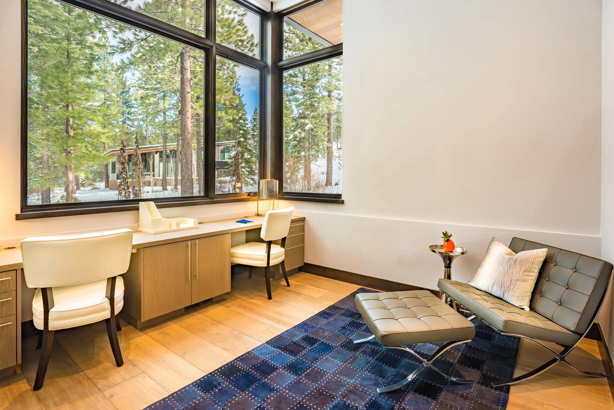Interior of a modern room with large window showing trees outside. Contains a white desk with two white chairs, a beige lounge chair with matching ottoman, a small side table with a decorative orange object and green plant, and a dark blue area rug.
