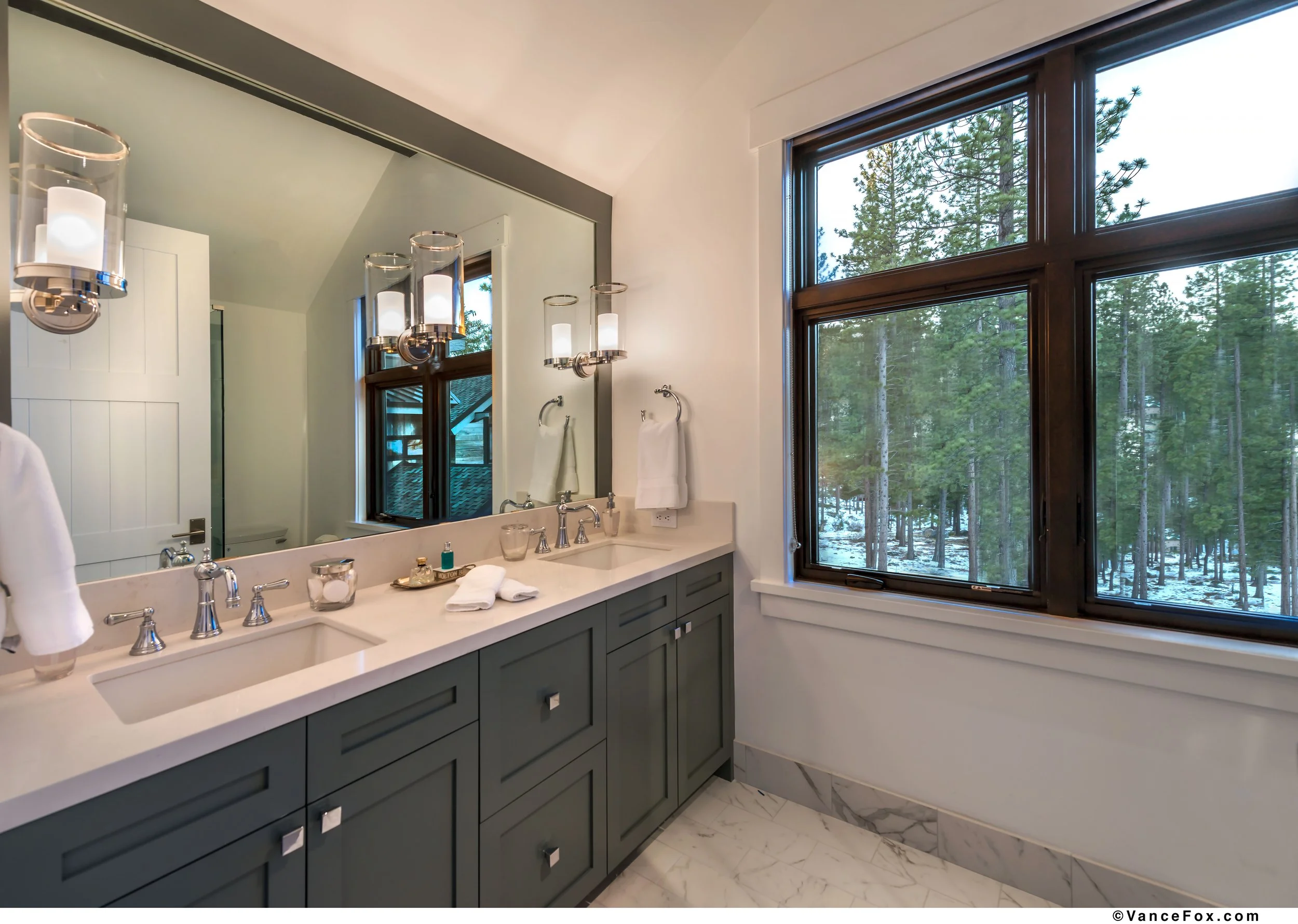 Bathroom with double vanity, large mirror, modern fixtures, and large window showing a wooded landscape outside.