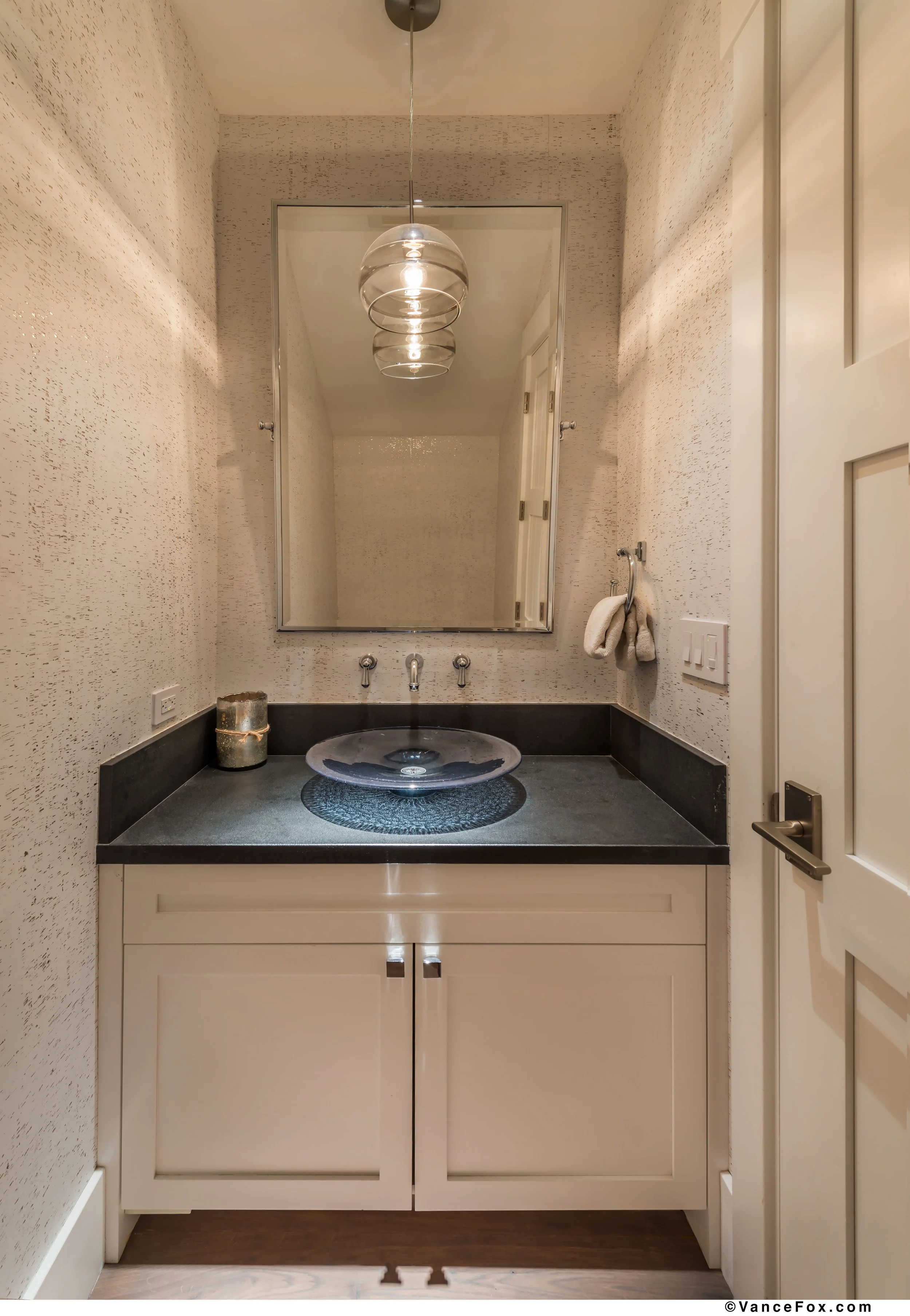 A small bathroom with white cork wallpapered walls, a black leathered granite countertop with a glass vessel sink, a large rectangular pivot mirror, a glass pendant light over the sink, and a white shaker cabinet. Custom stained European Oak floors.