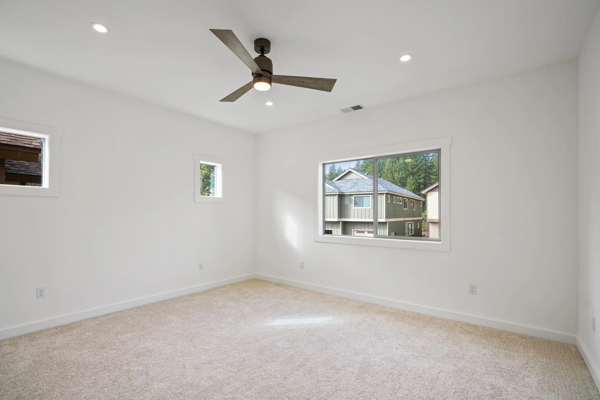 Empty room with white walls, beige carpet, large window, smaller windows, ceiling fan, and recessed lighting.