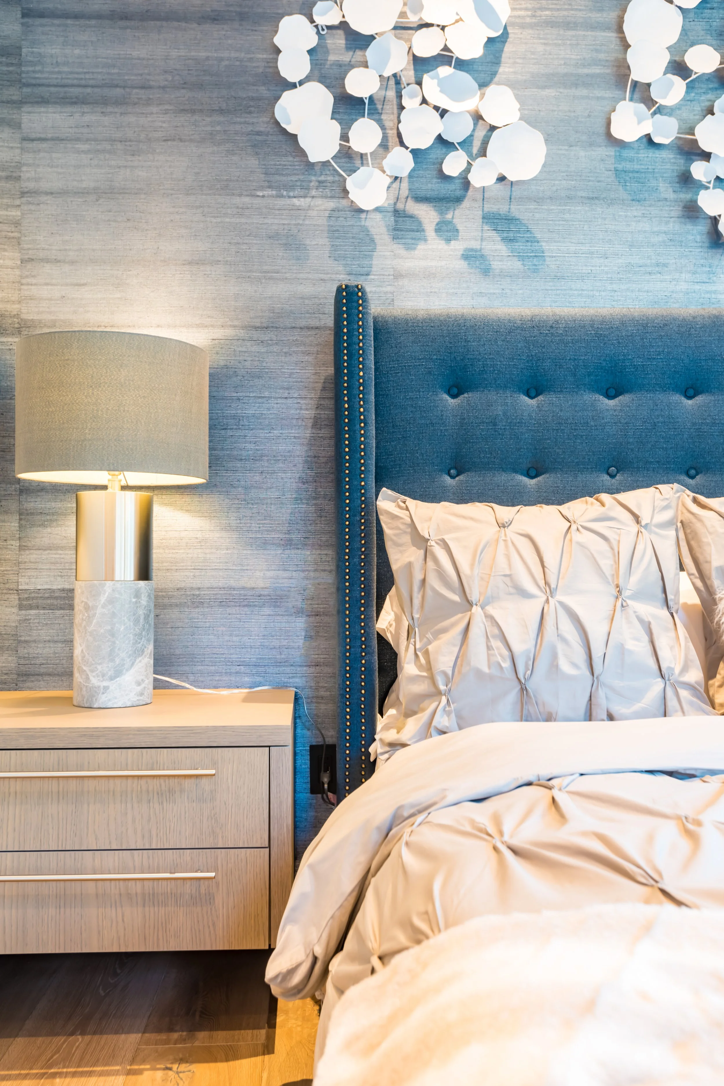 A bedroom with beige bedding, a blue upholstered headboard with nailhead trim, a nightstand with a textured marble lamp, and decorative white wall art resembling folded paper floral designs.