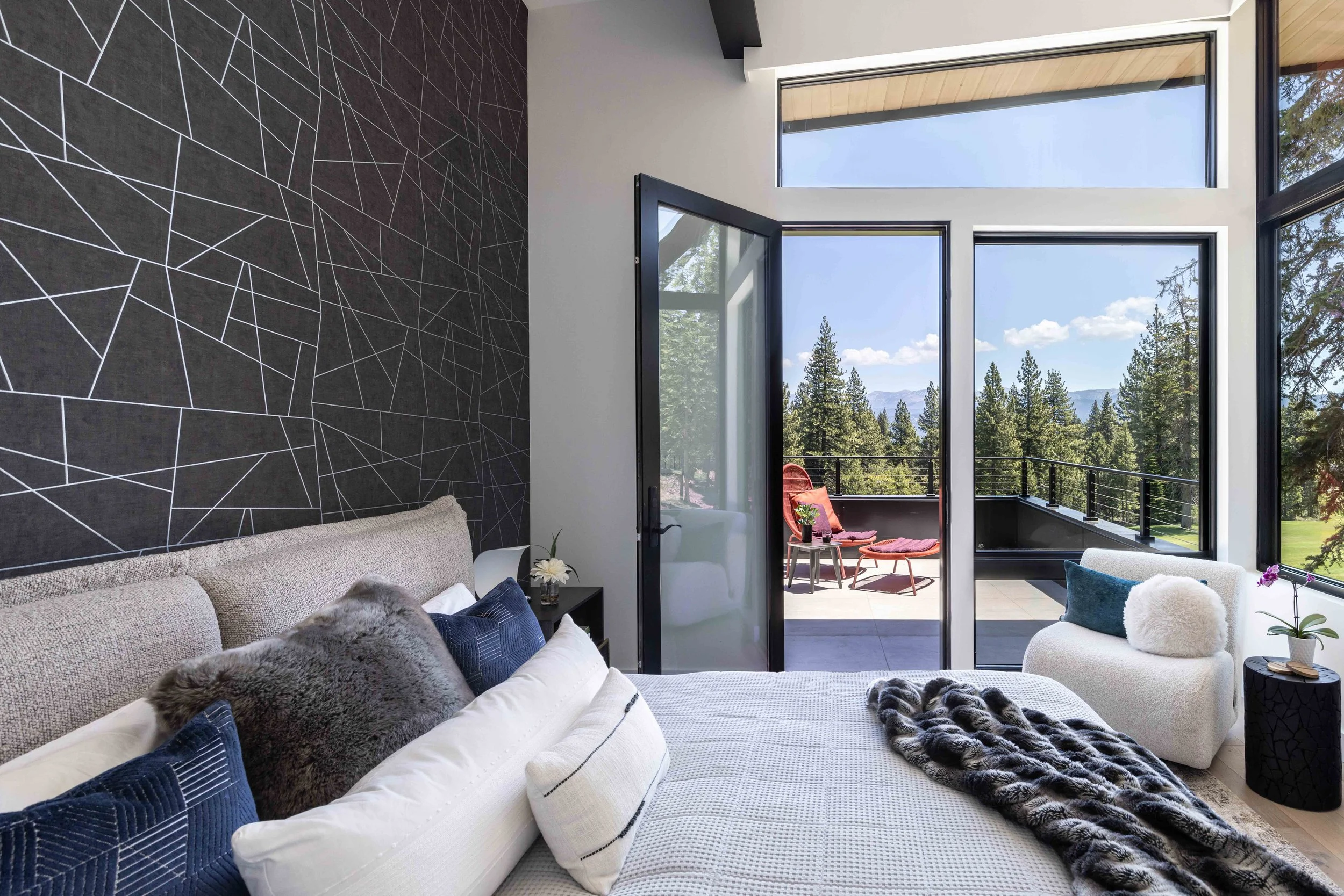 Modern bedroom with large glass windows and balcony overlooking a forest with mountains in the distance.