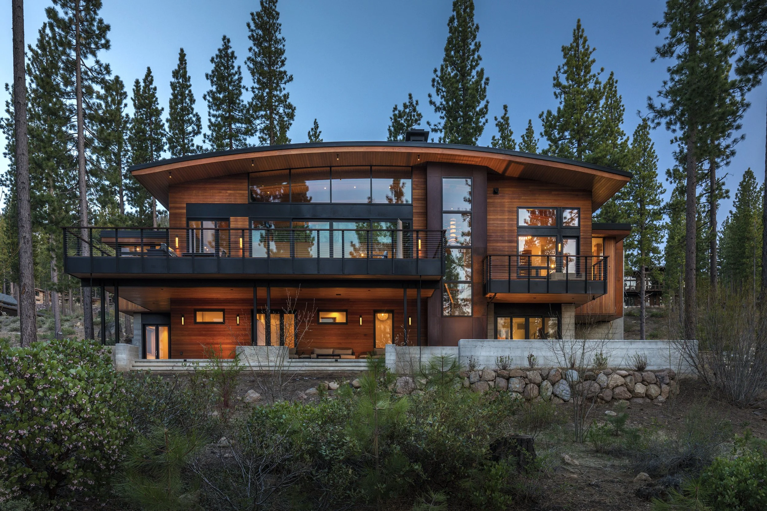 Modern two-story house with large glass windows and balconies, surrounded by trees, during dusk.