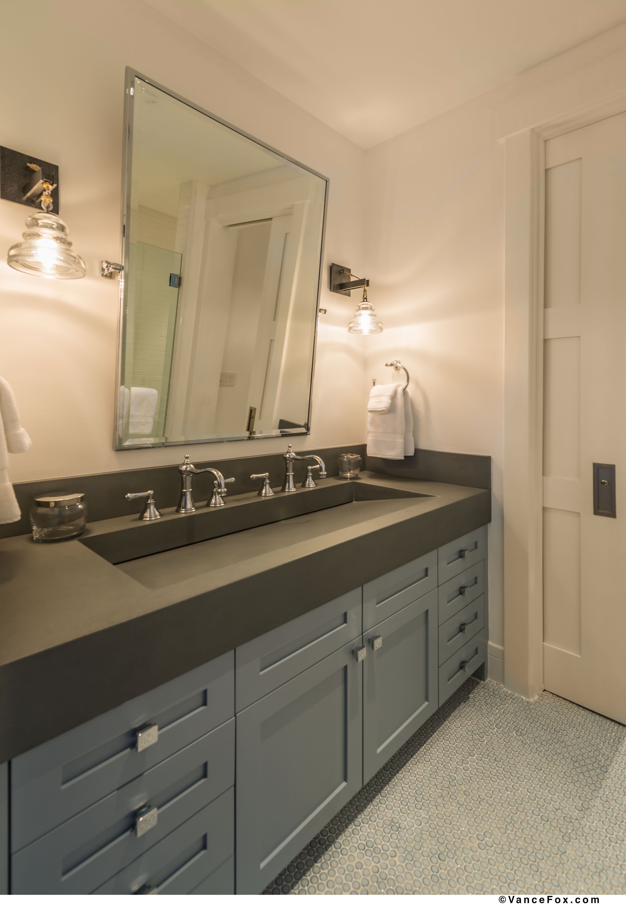 A bathroom with a gray vanity, a large mirror, and wall-mounted light fixtures. The vanity has multiple drawers and cabinets, with a dark countertop and a built-in sink with silver faucets. There are white towels hanging on the wall and a small glass