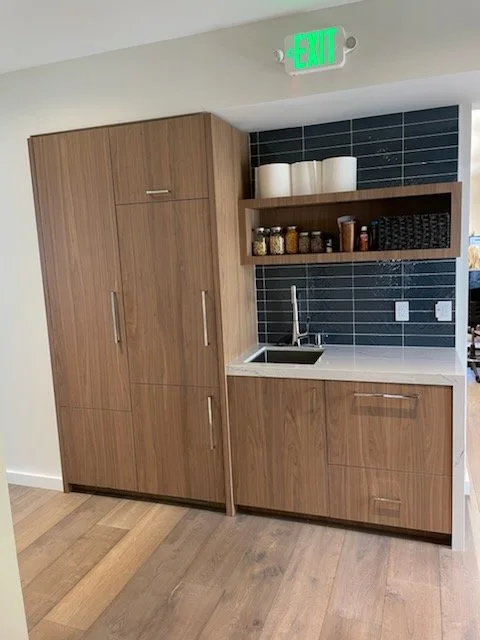 A small kitchenette with wooden cabinets, a black tiled backsplash, a white countertop with a sink, three white canisters on top, and a shelf with spice jars, set against a wall with black tiles. An emergency exit sign is above.