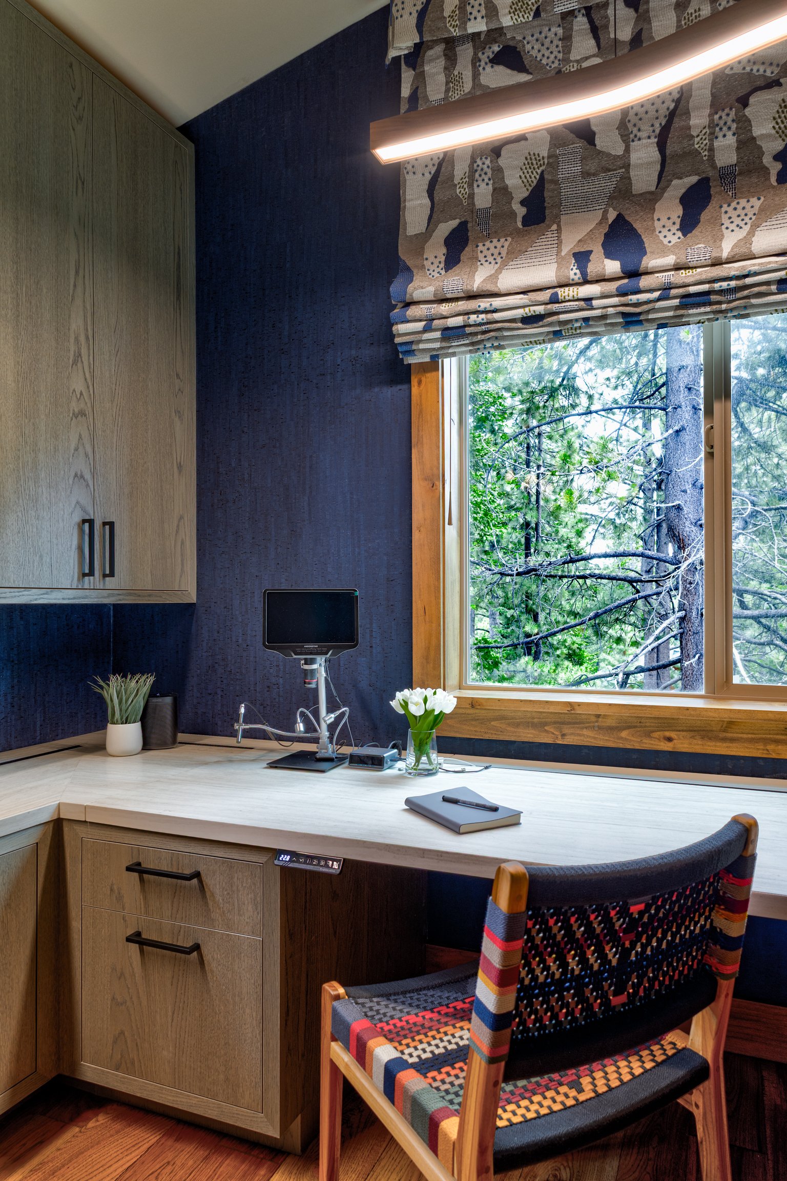 Home office with a wooden desk, colorful woven chair, black wall, window with greenery outside, small monitor, potted plants, vase with white flowers, notebook with pen, and a modern light fixture.