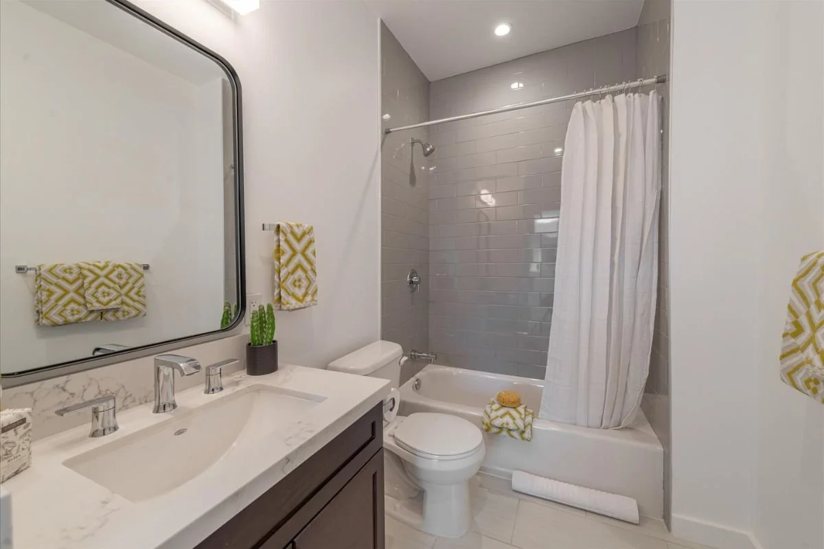 Modern bathroom with a white vanity, marble countertop, and a large mirror. There are yellow and white patterned towels, a small potted cactus, a toilet, and a bathtub with a shower. The shower has gray tiles and a white curtain.