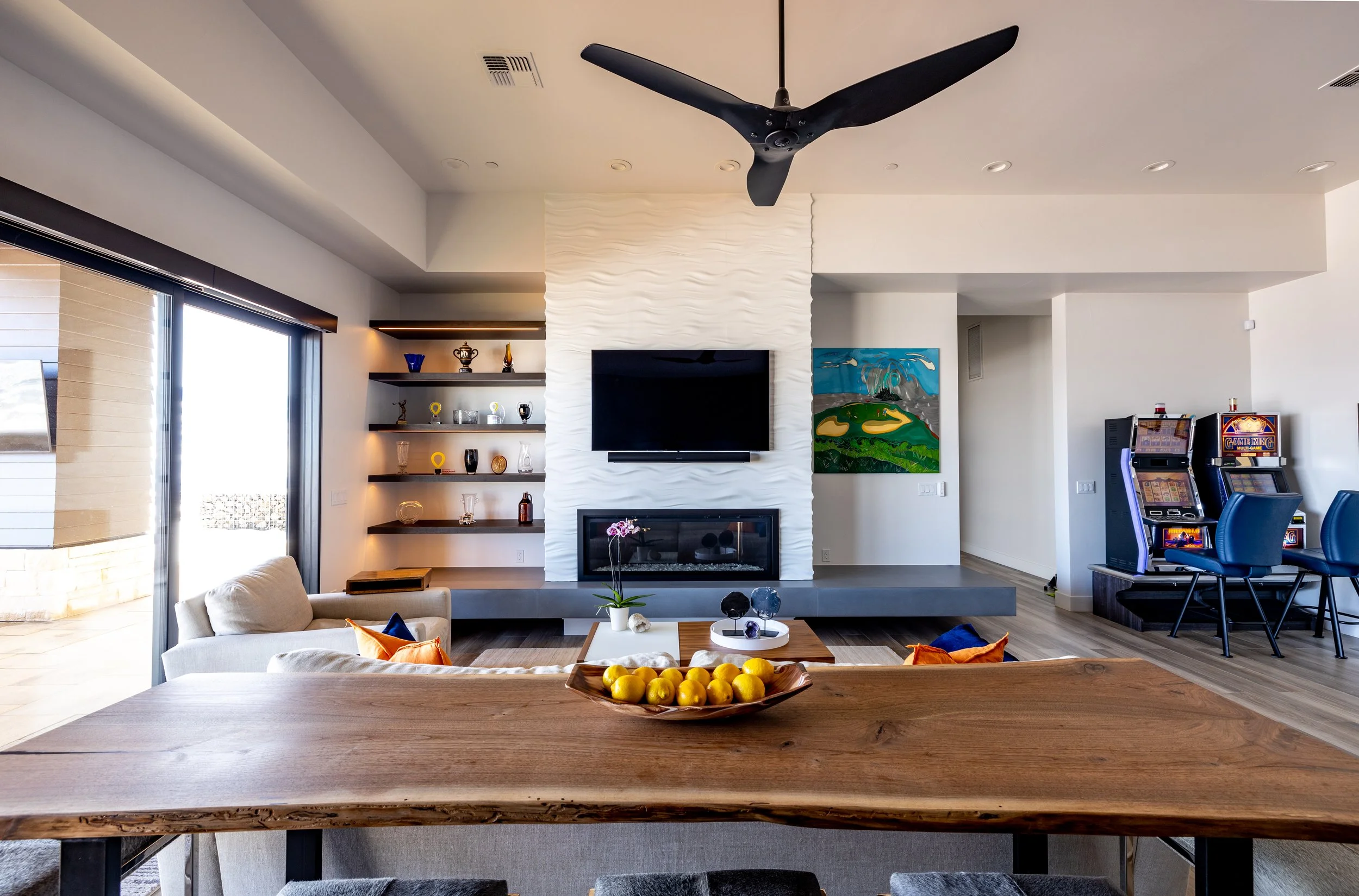Modern living room with a white textured wall, a mounted TV above a fireplace, a sitting area with colorful pillows, a wooden dining table with lemons, and a window with sliding glass doors leading outside. There are built-in shelves with decorative 