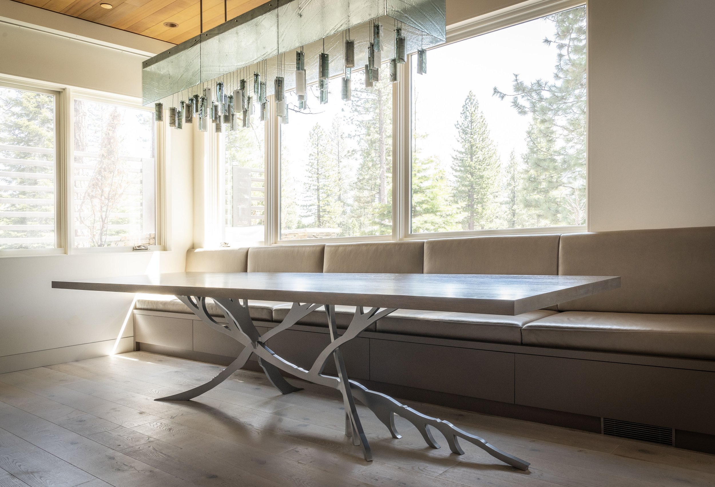 Modern dining area with a long wooden table featuring an intricate metal base, beige cushioned bench seating along large windows with views of trees, and a geometric chandelier hanging from the ceiling.