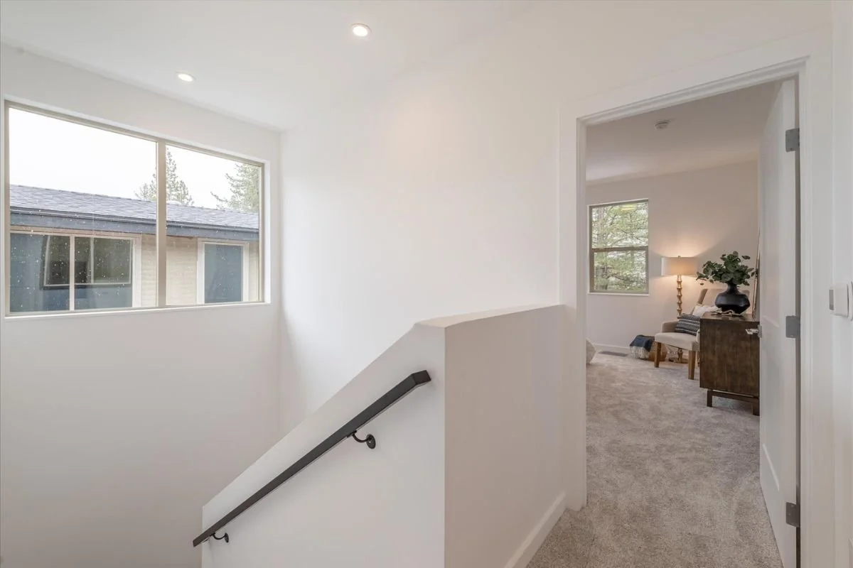 A bright residential hallway with large window, beige carpet, and an open doorway leading to a living room with a lamp and chair.