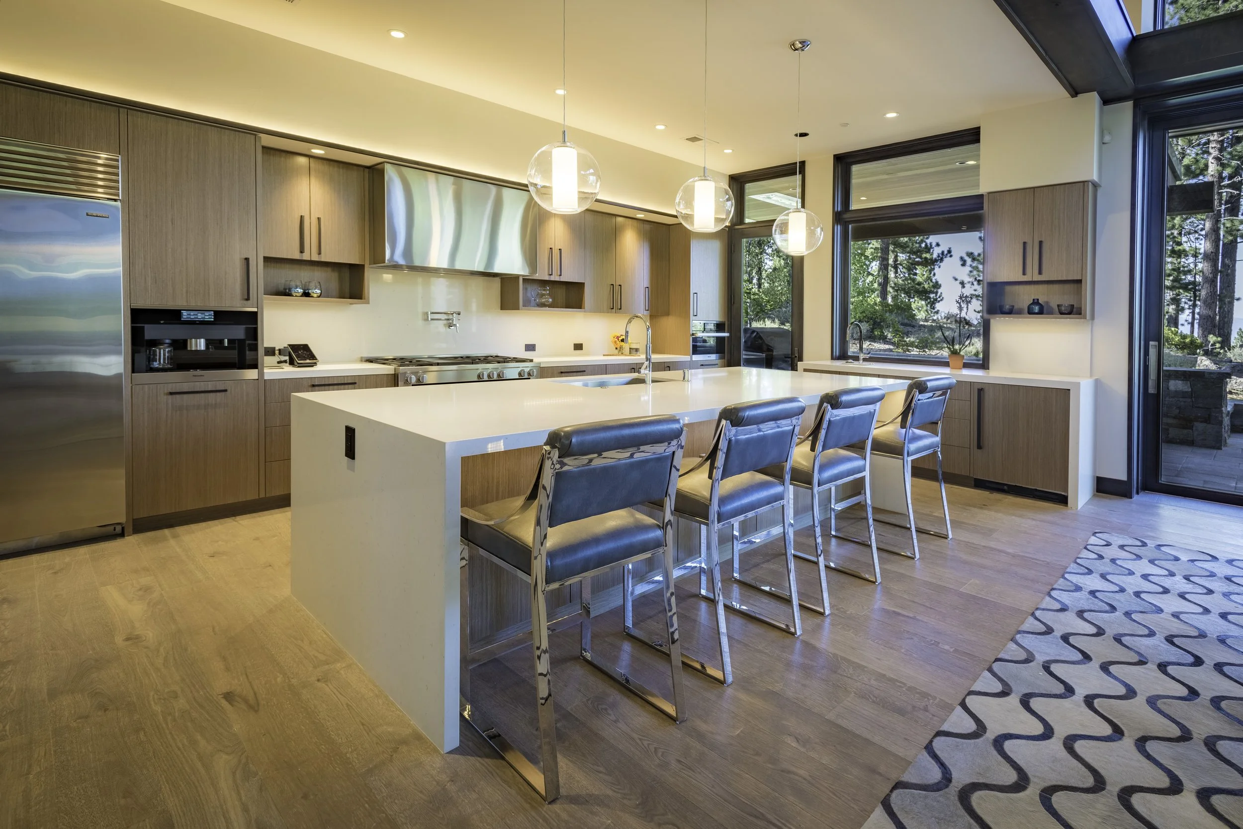 Modern kitchen with wooden cabinets, white island, stainless steel appliances, pendant lighting, large windows, and hardwood flooring.