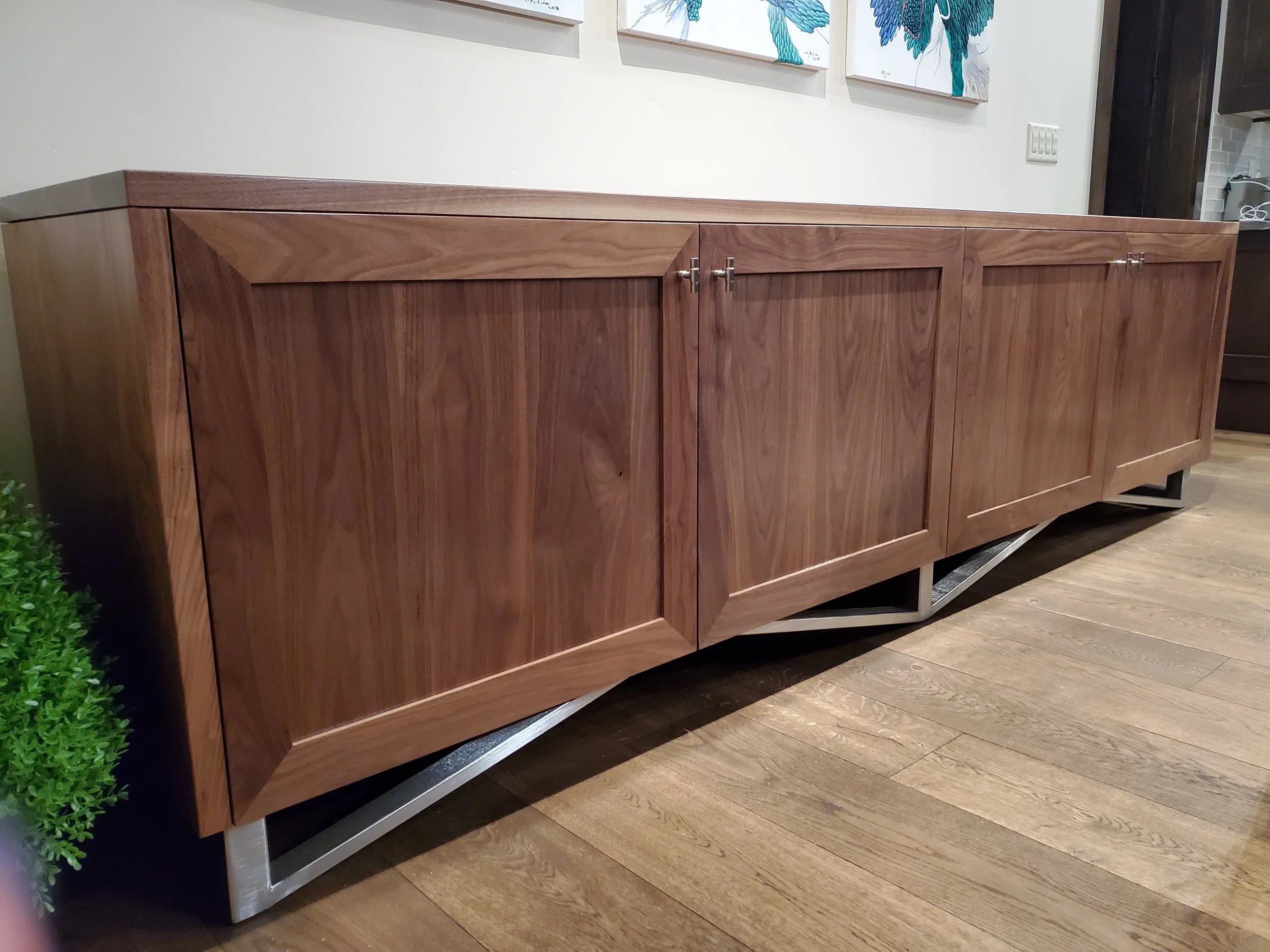 A wooden sideboard with four panel doors and metal handles on a wooden floor, with artwork above it and a small green plant at the left edge.
