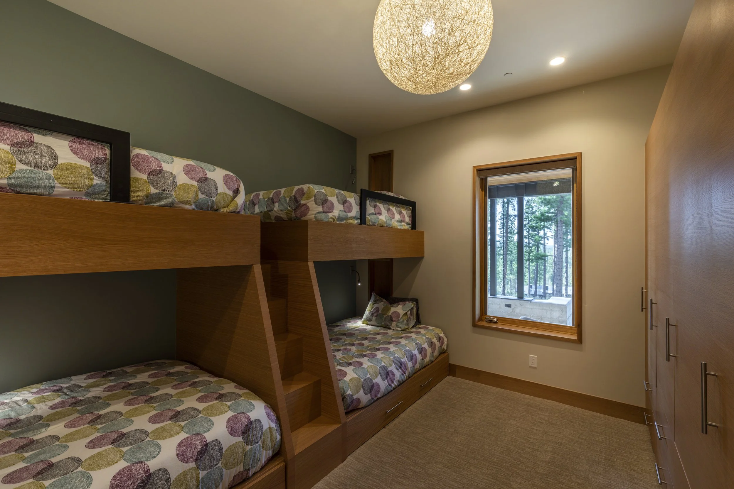 A bedroom with a bunk bed, featuring colorful dotted bedding, a window with a view of trees, a wooden wardrobe, and a textured ceiling light.
