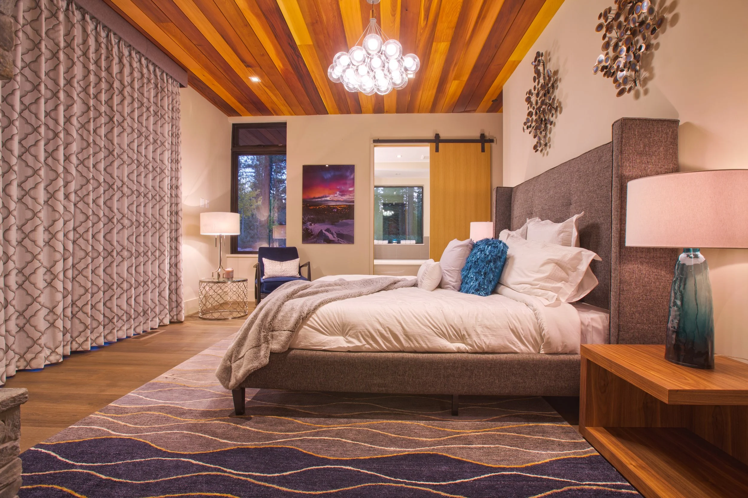A bedroom with a wooden ceiling, a large bed with white bedding and decorative pillows, a nightstand with a lamp, a window, a chair, and a sliding barn door.