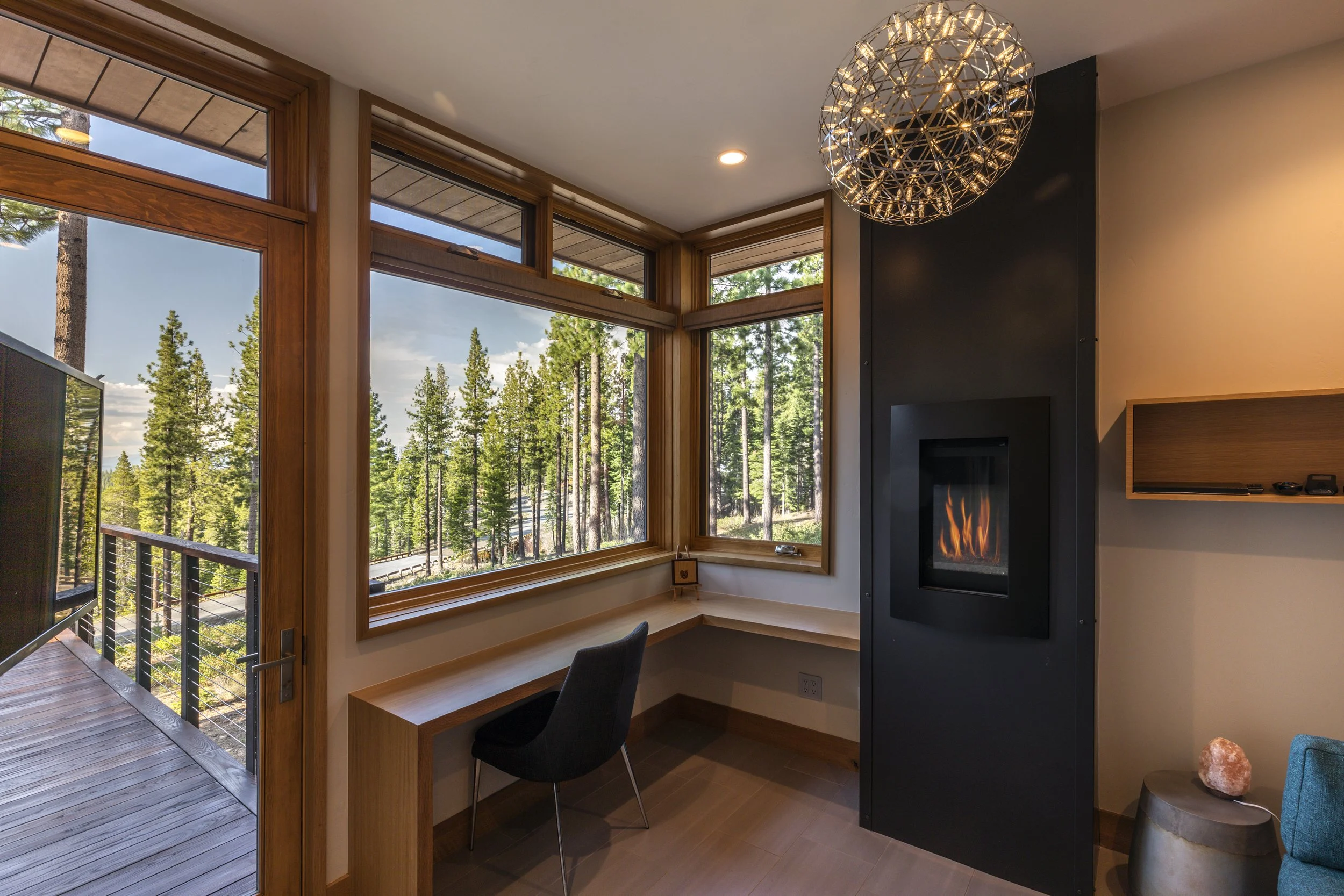 Interior of a room with large windows overlooking a forest, a built-in wooden desk, a black office chair, a modern fireplace, a hanging spherical light fixture, and a small side table with a salt lamp.