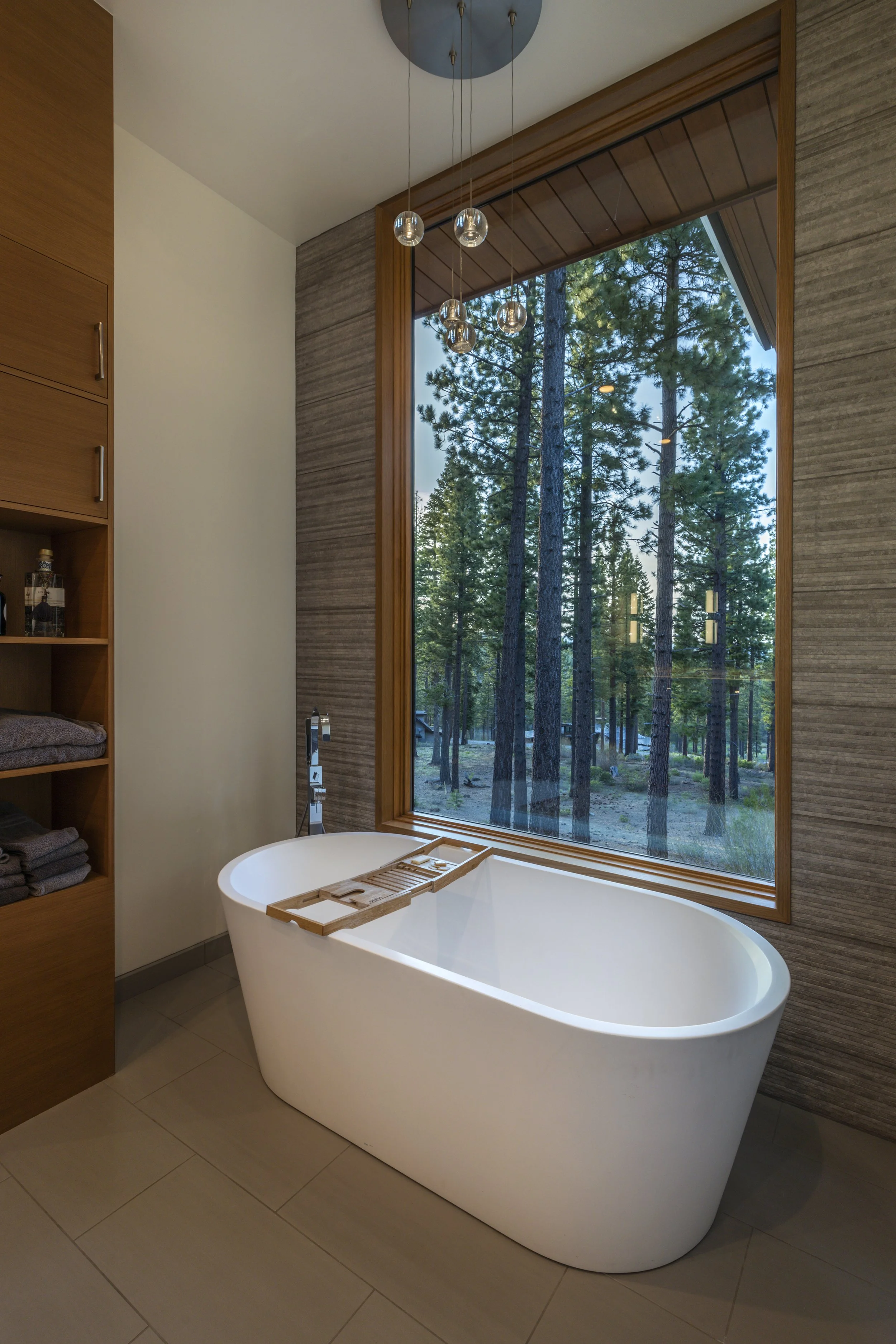 Modern bathroom with a white freestanding bathtub placed near a large window overlooking a forest of tall pine trees. Wooden elements frame the window and a wooden bath tray is placed across the tub. A contemporary light fixture hangs from the ceilin