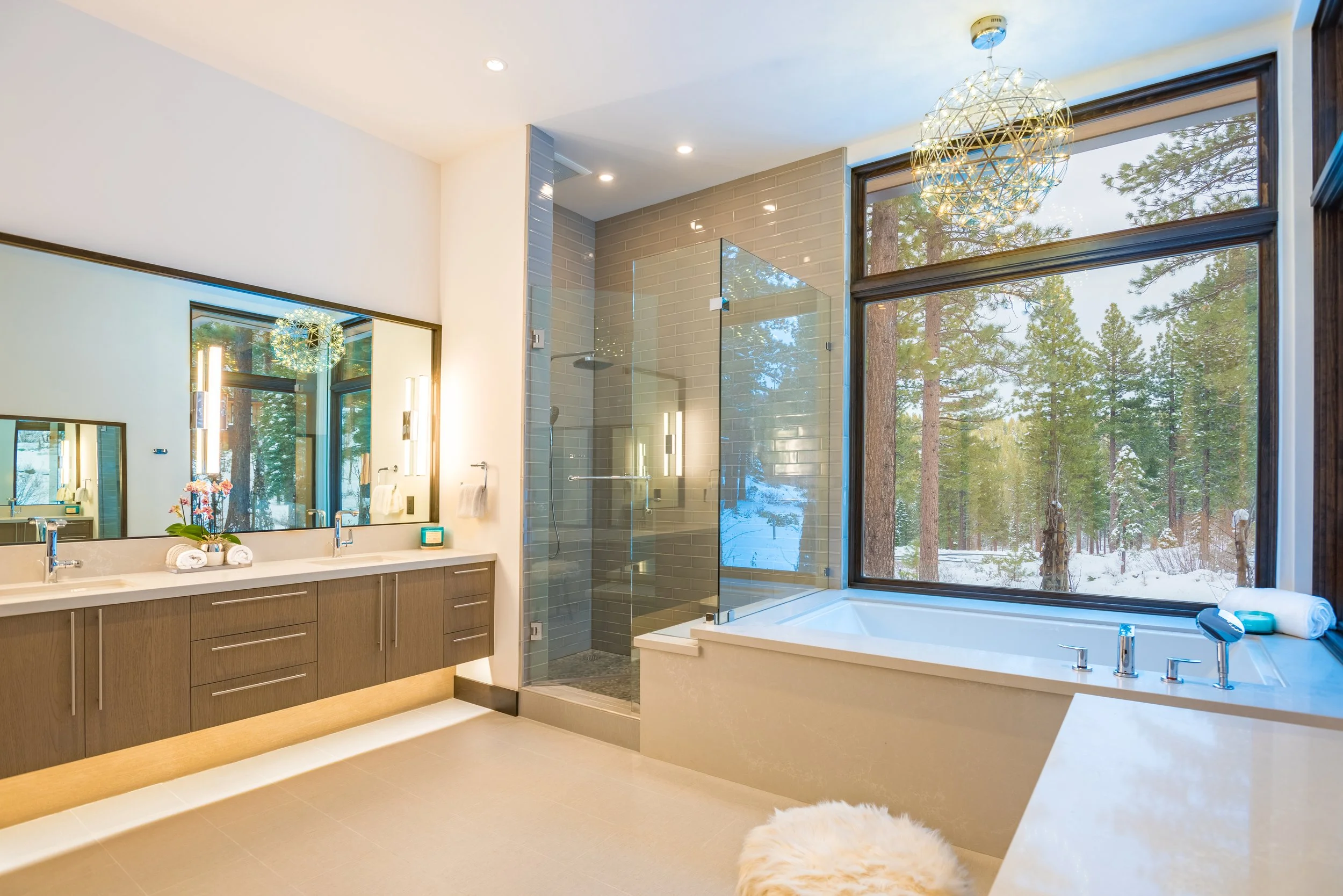 Modern bathroom with large window showing snowy forest outside; includes double vanity with mirror, shower with glass enclosure, and bathtub beside window.
