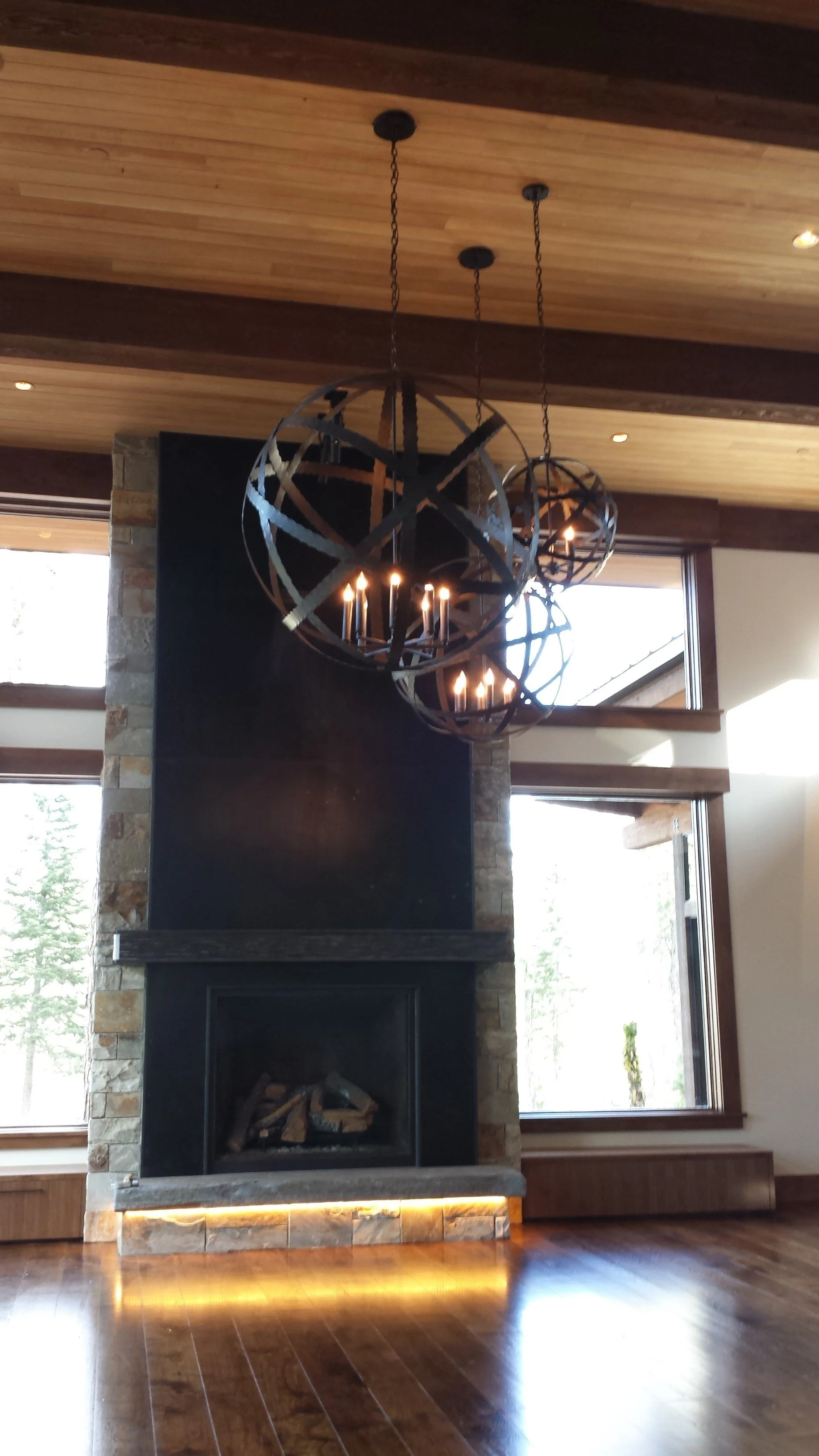 Modern living room fireplace with a black surround and stacked stone facade, large windows on the sides, and a wooden ceiling with custom designed metal sphere chandeliers by Emily Roose.