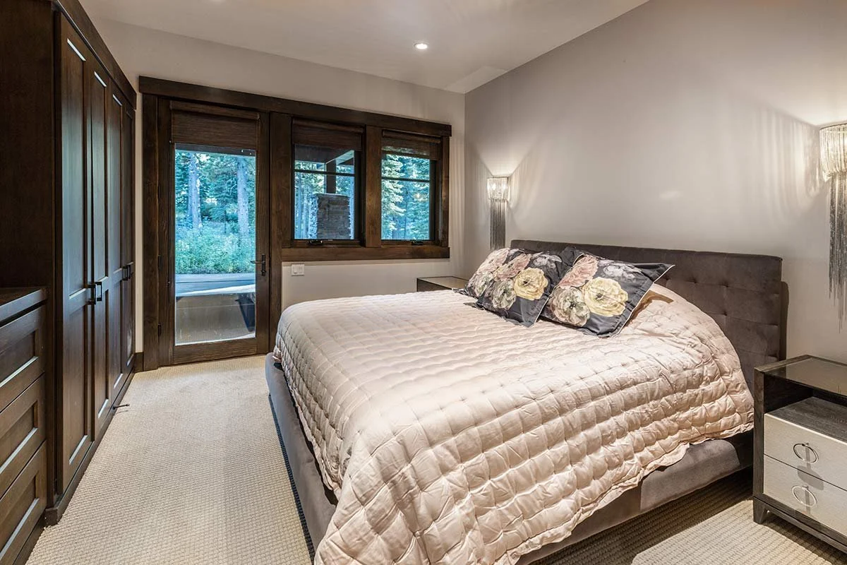 A bedroom with a large bed, dark wooden wardrobe, a nightstand, and windows showing a wooded outdoor scene.