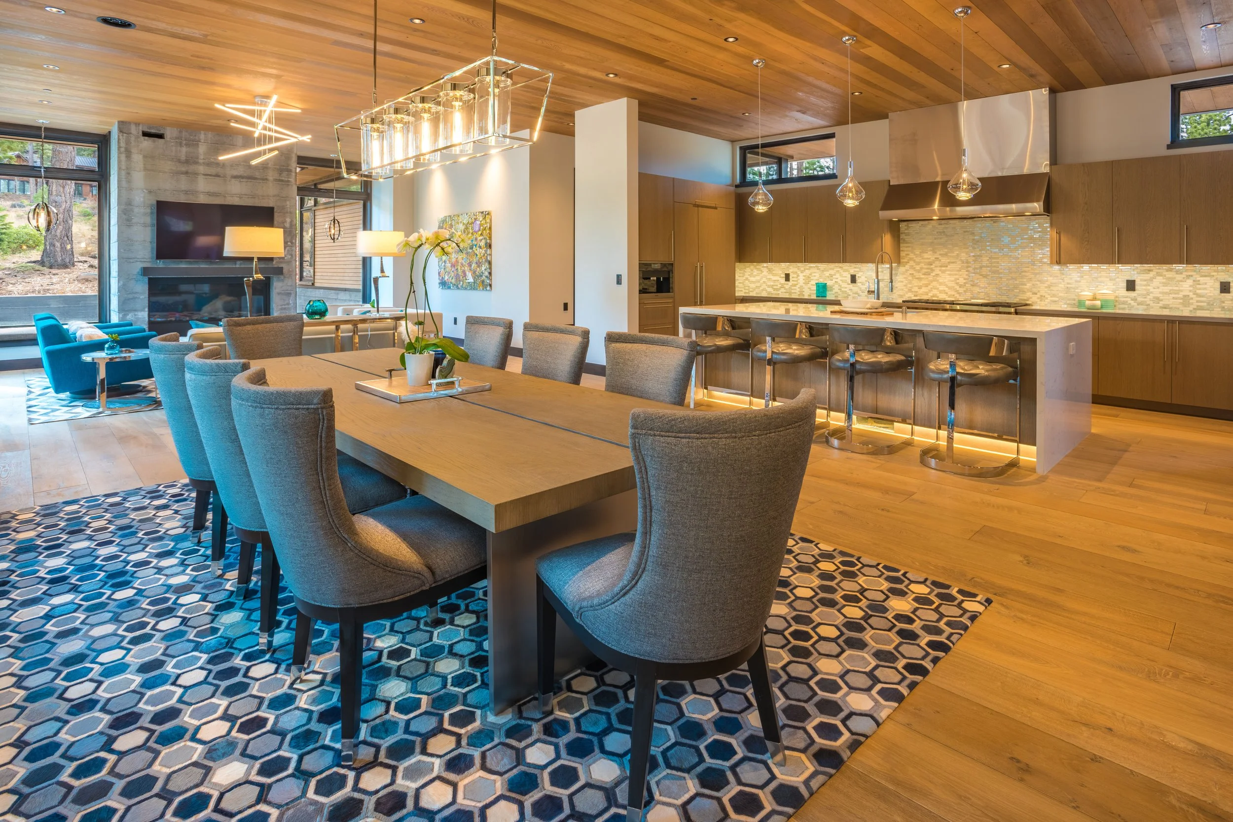 An open-concept living and kitchen area with a large wooden dining table, upholstered dining chairs, a patterned rug, a modern kitchen with an island and bar stools, wood cabinets, and a wood-paneled ceiling, along with windows showing outdoor greene