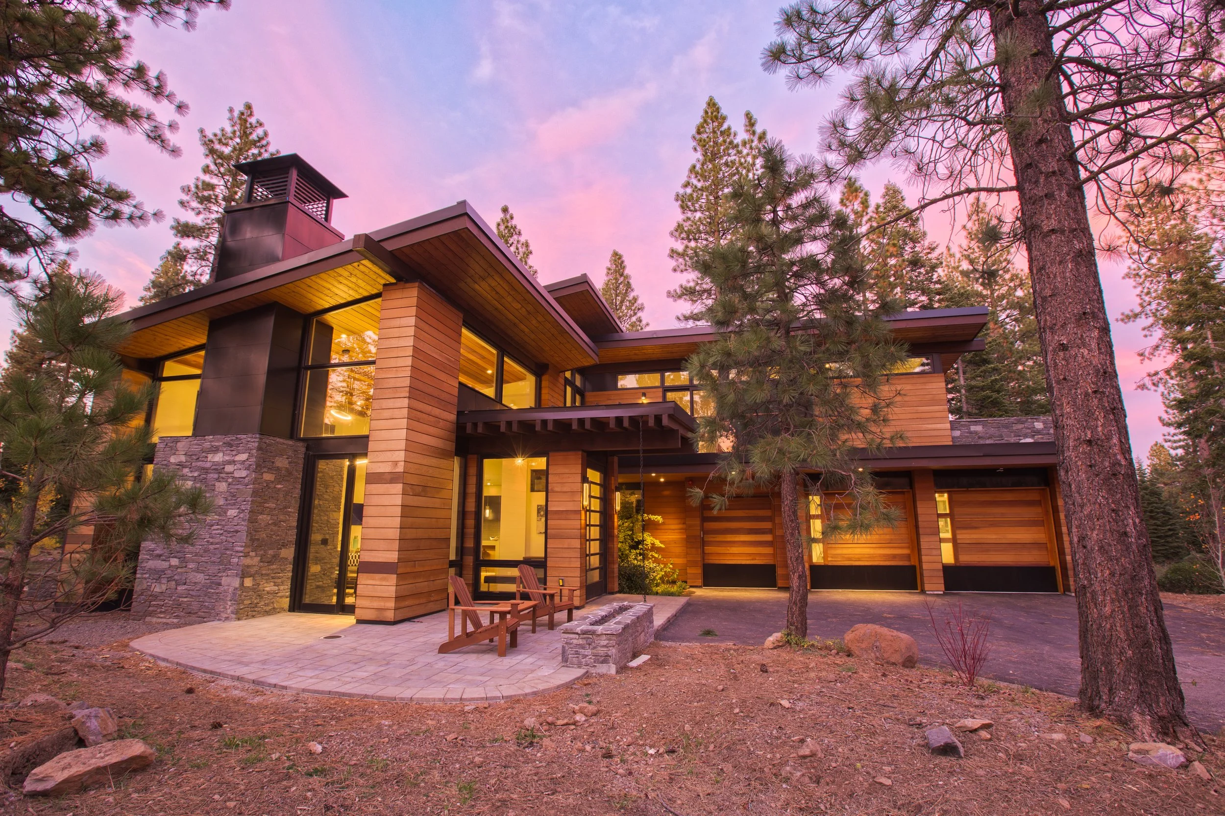 Modern house with large windows, wooden and stone exterior, situated among tall pine trees during sunset.