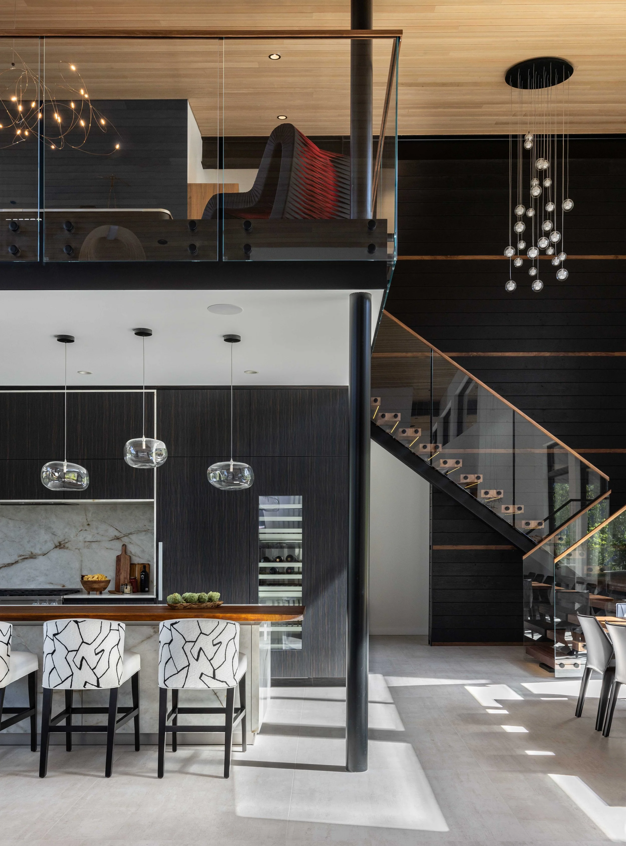 Modern kitchen and dining area with black and white decor, featuring a kitchen island with bar stools, a staircase with glass railing, and stylish pendant and chandelier lighting.