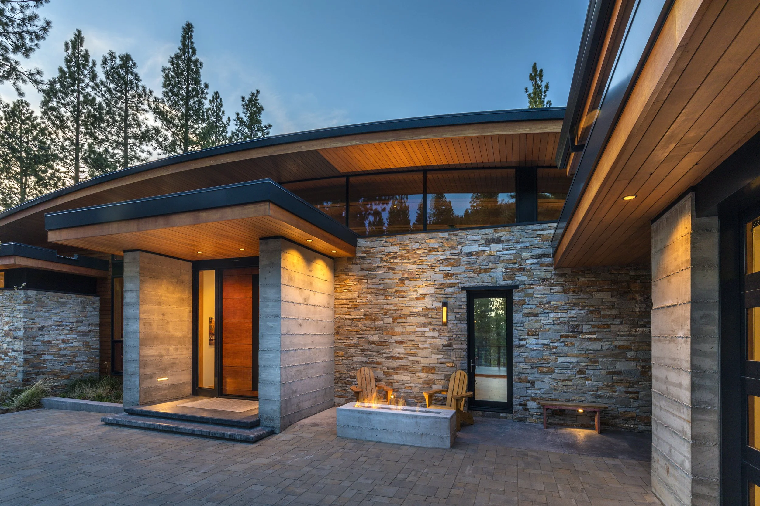 Modern house exterior with stone and wood walls, large glass windows, outdoor seating around a fire pit, surrounded by tall pine trees at dusk.