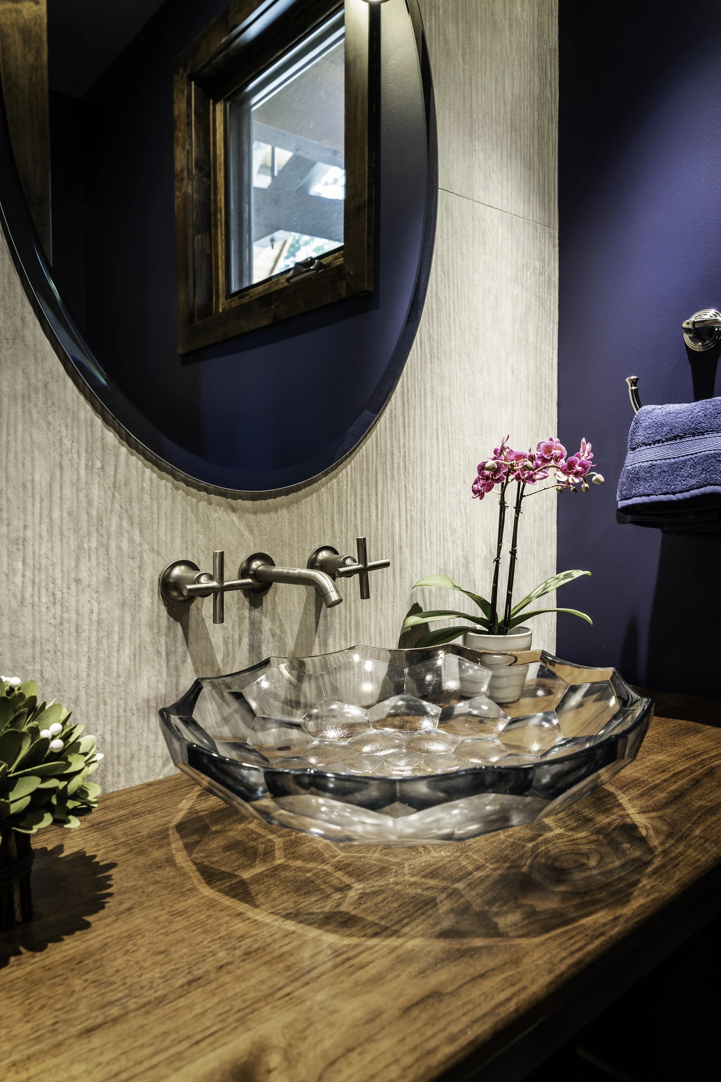 Close-up of a bathroom sink area with a wooden countertop, decorative glass bowl, and pink orchid plant in a white pot. The wall features a large round mirror, a small wooden window, and blue and beige paint.