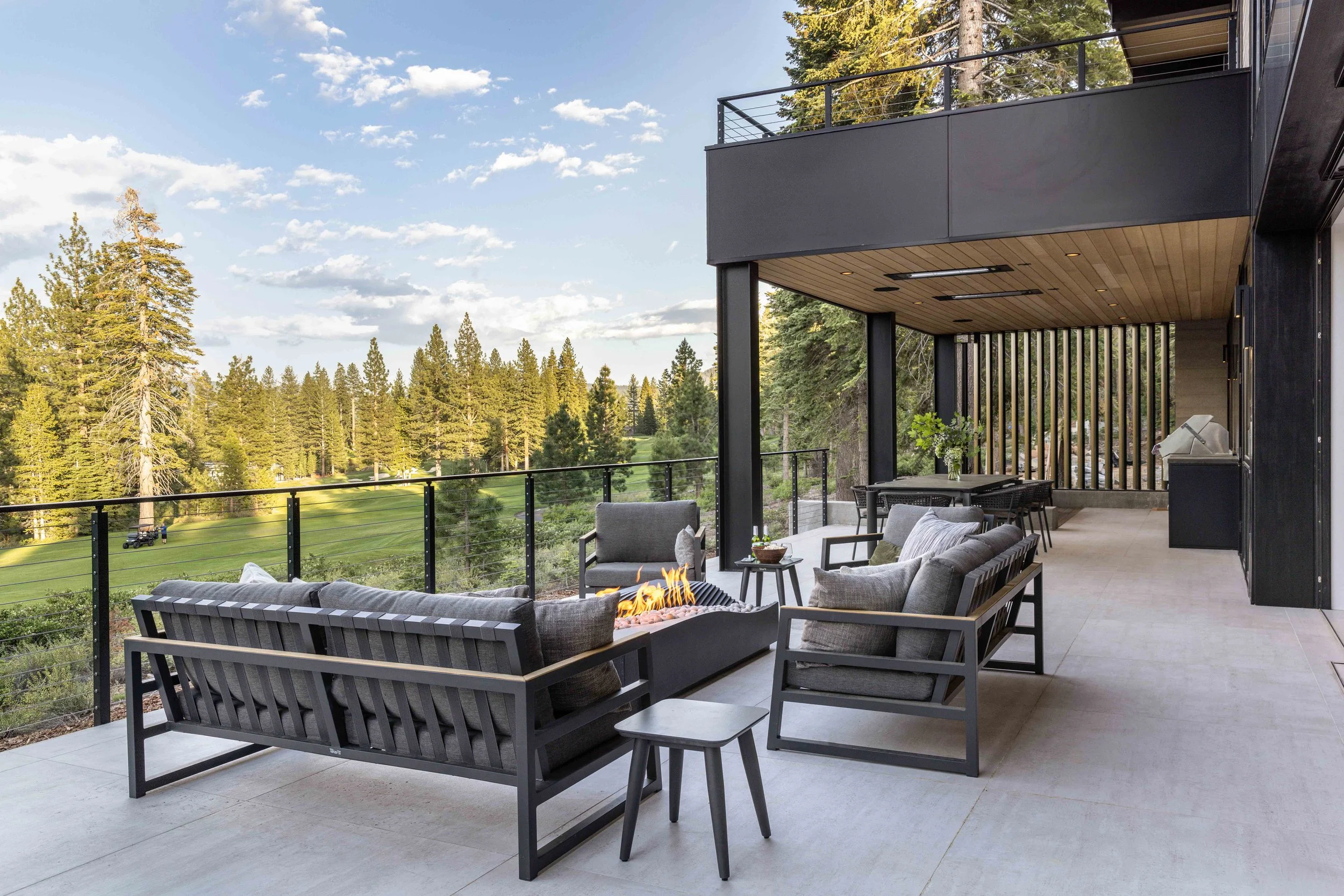 Modern outdoor balcony with seating area, fireplace, and scenic view of pine trees and a golf course.