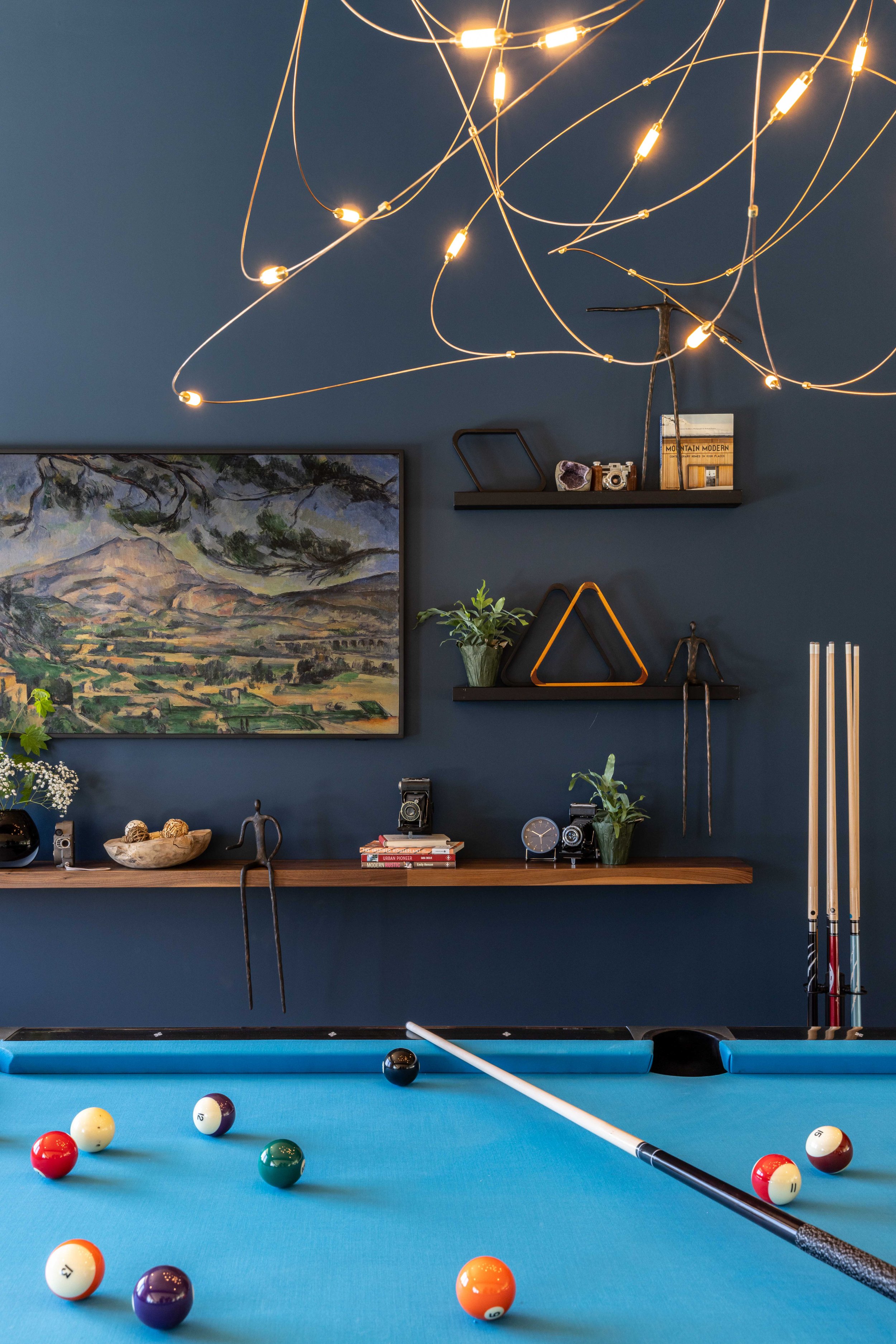 A teal pool table with billiard balls and cue stick. Behind, a dark blue wall with a large landscape painting, a floating shelf with books and decorative objects, including plants, cameras, and sculptures. Overhead, a modern chandelier with multiple 
