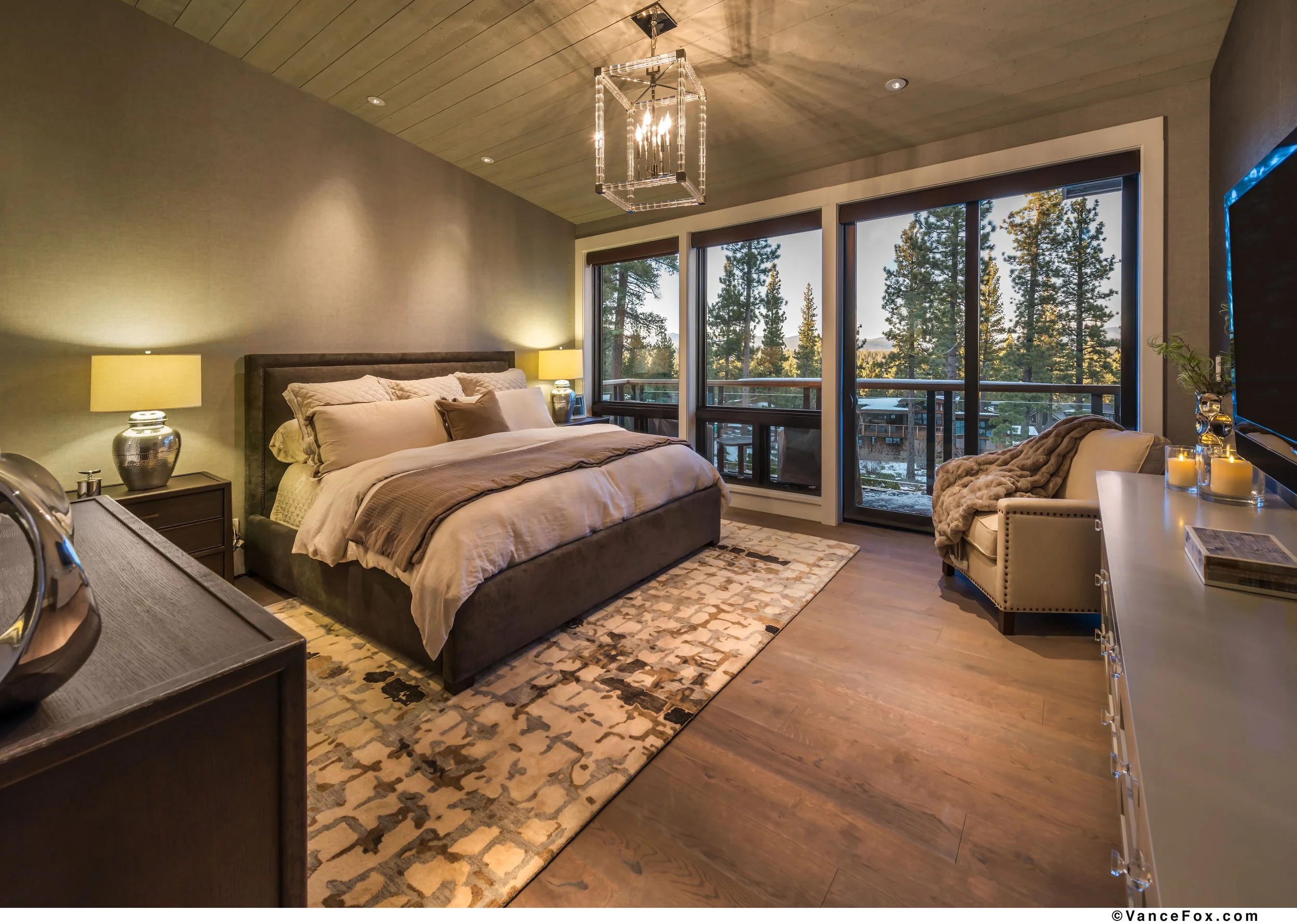 A cozy junior primary bedroom bedroom with a large bed, nightstands with lamps, a patterned area rug, large windows with a view of trees, a sitting area with a chair and side table, and elegant lighting.
