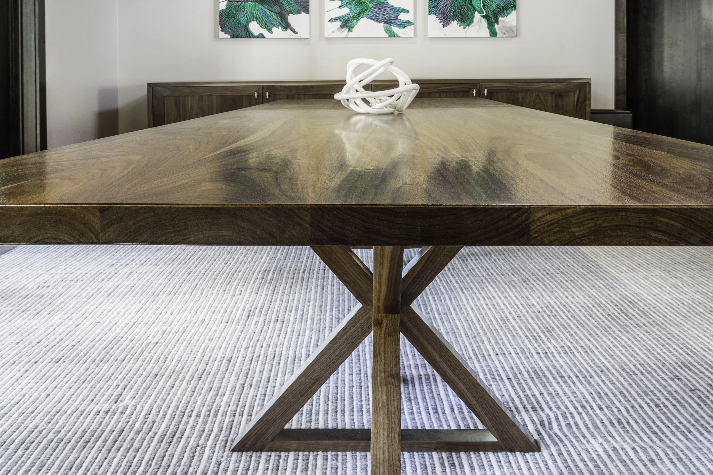 A wooden dining table with a white decorative sculpture in the center, placed on a white striped rug. In the background, there is a wall with three abstract green and blue paintings.
