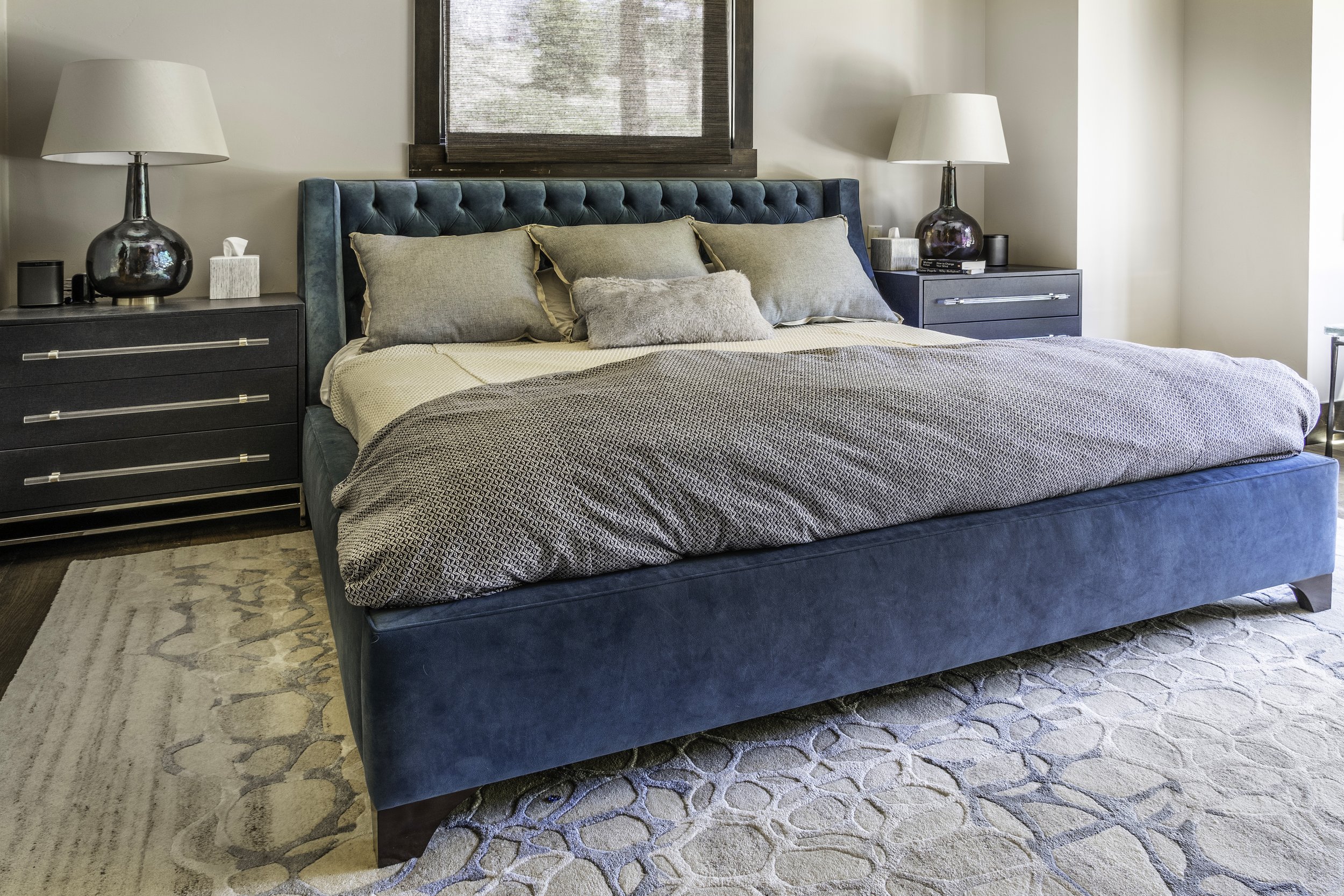 A bedroom with a blue upholstered bed, beige pillows, dark nightstands on each side, lamps, a window with a brown blind, and a patterned area rug.