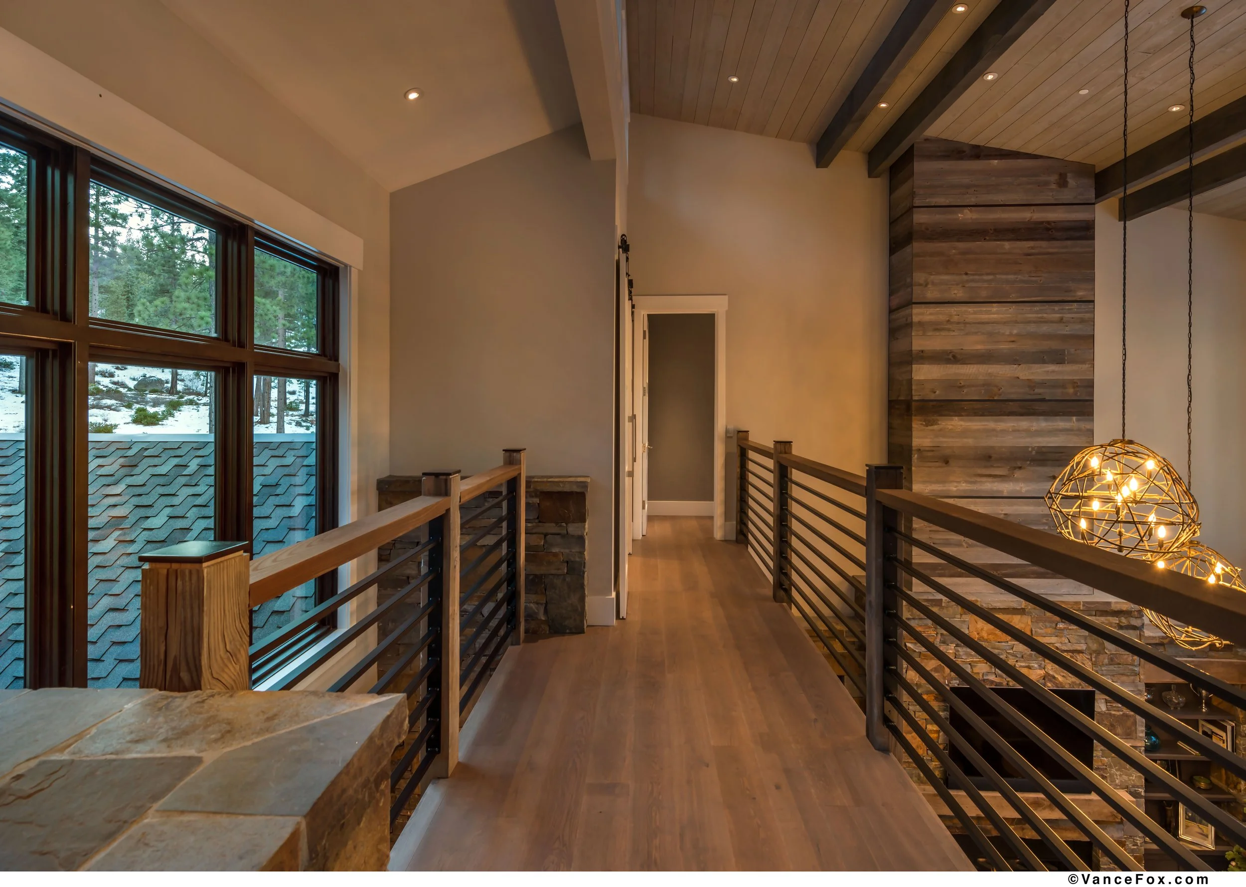 Interior view of a modern home loft with wooden flooring, large windows showing a snowy landscape, a stone and wood accent wall, and contemporary pendant lighting.