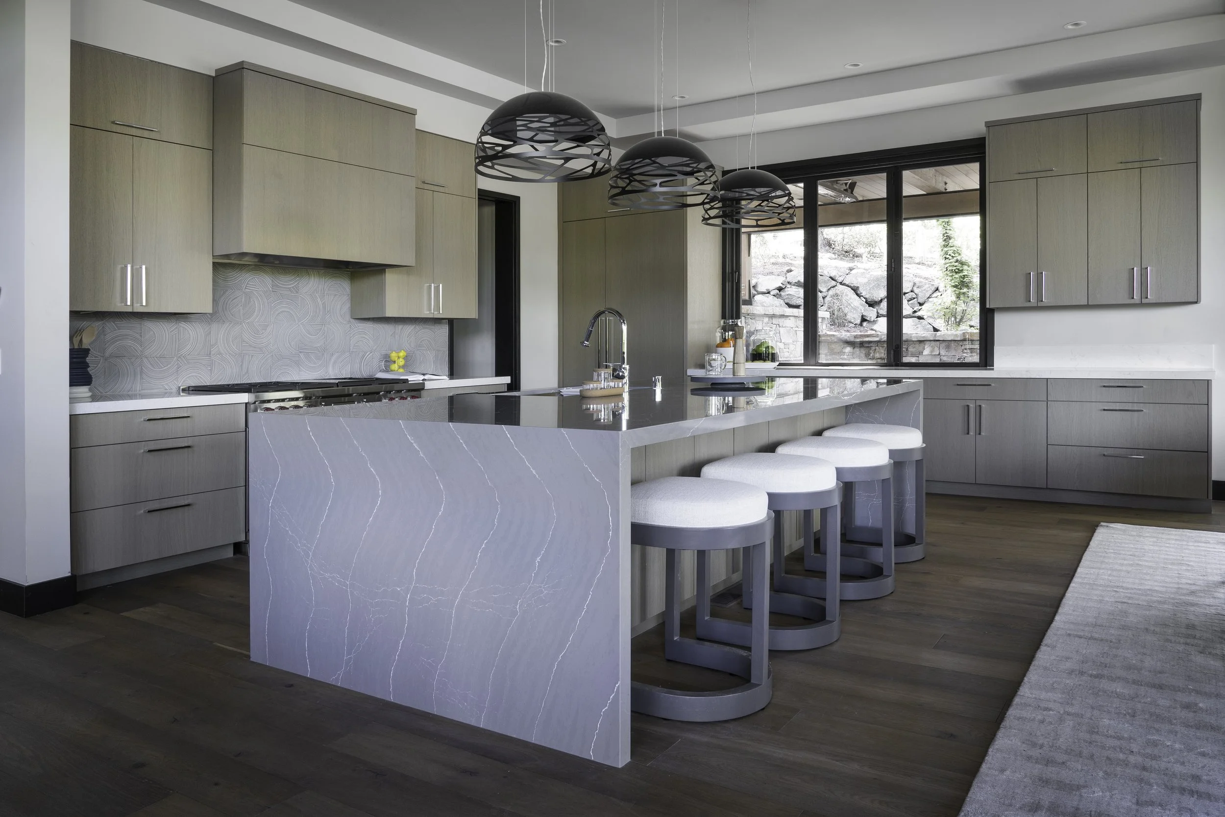 Modern kitchen with light wood cabinets, white countertops, and a large island with bar stools. Black pendant lights hang above the island and a window overlooks a rocky outdoor landscape.