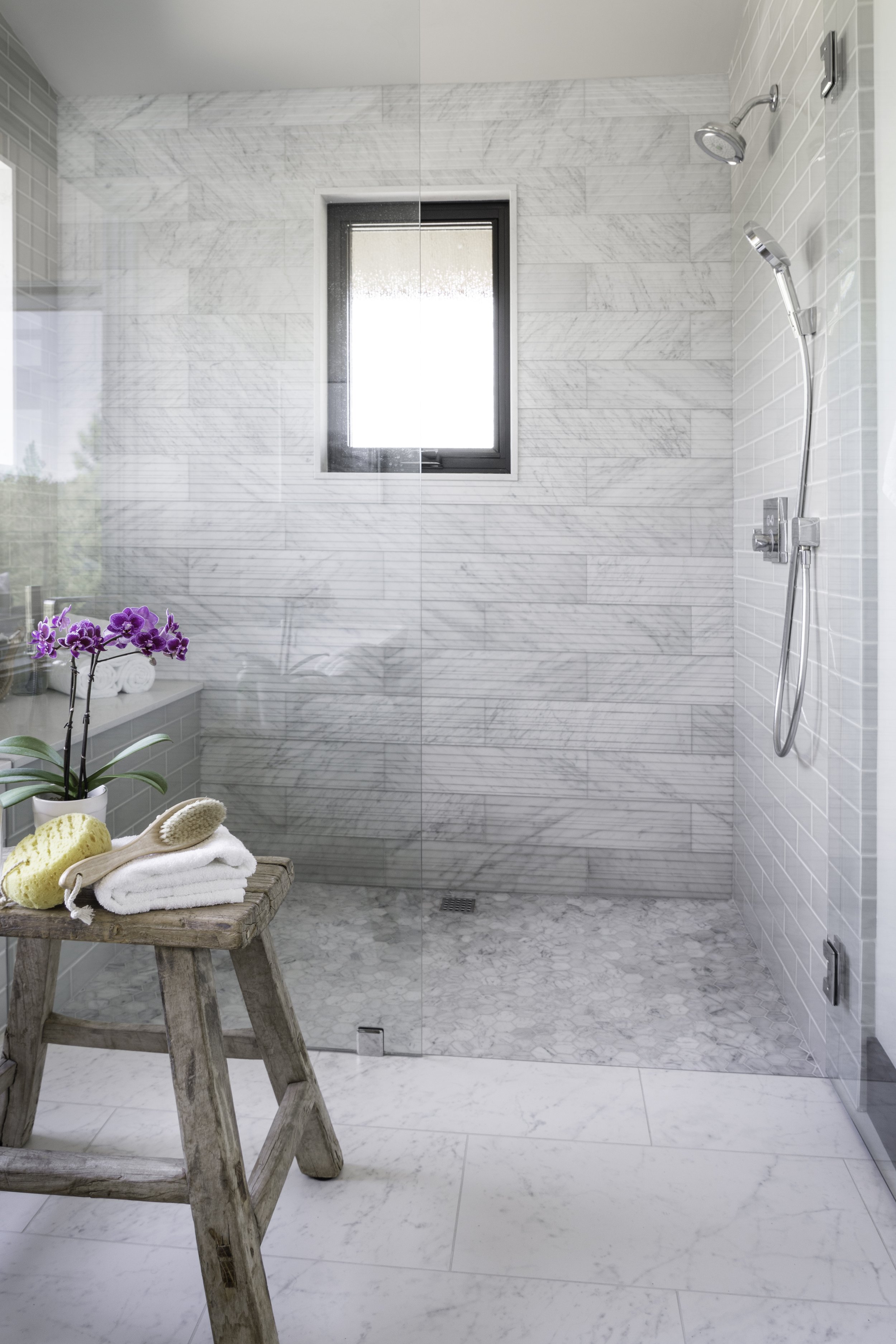 A modern walk-in shower with white marble tile walls and floors, a frosted window, and a glass door, with bathing accessories and purple orchids on a wooden stool.