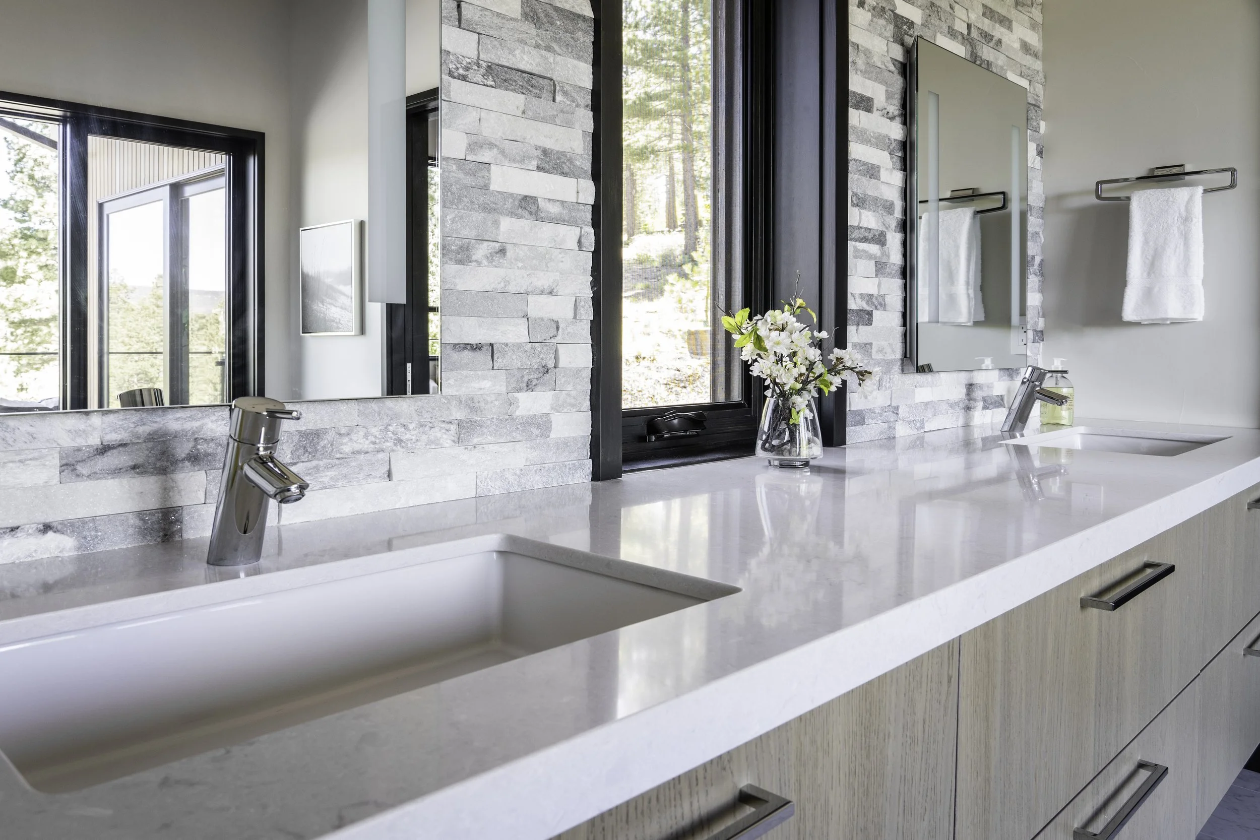 Modern bathroom double vanity with white countertop, two sinks, and chrome faucets, featuring a large mirror, a window with a view of trees, a vase with flowers, and white towels hanging on a rack.