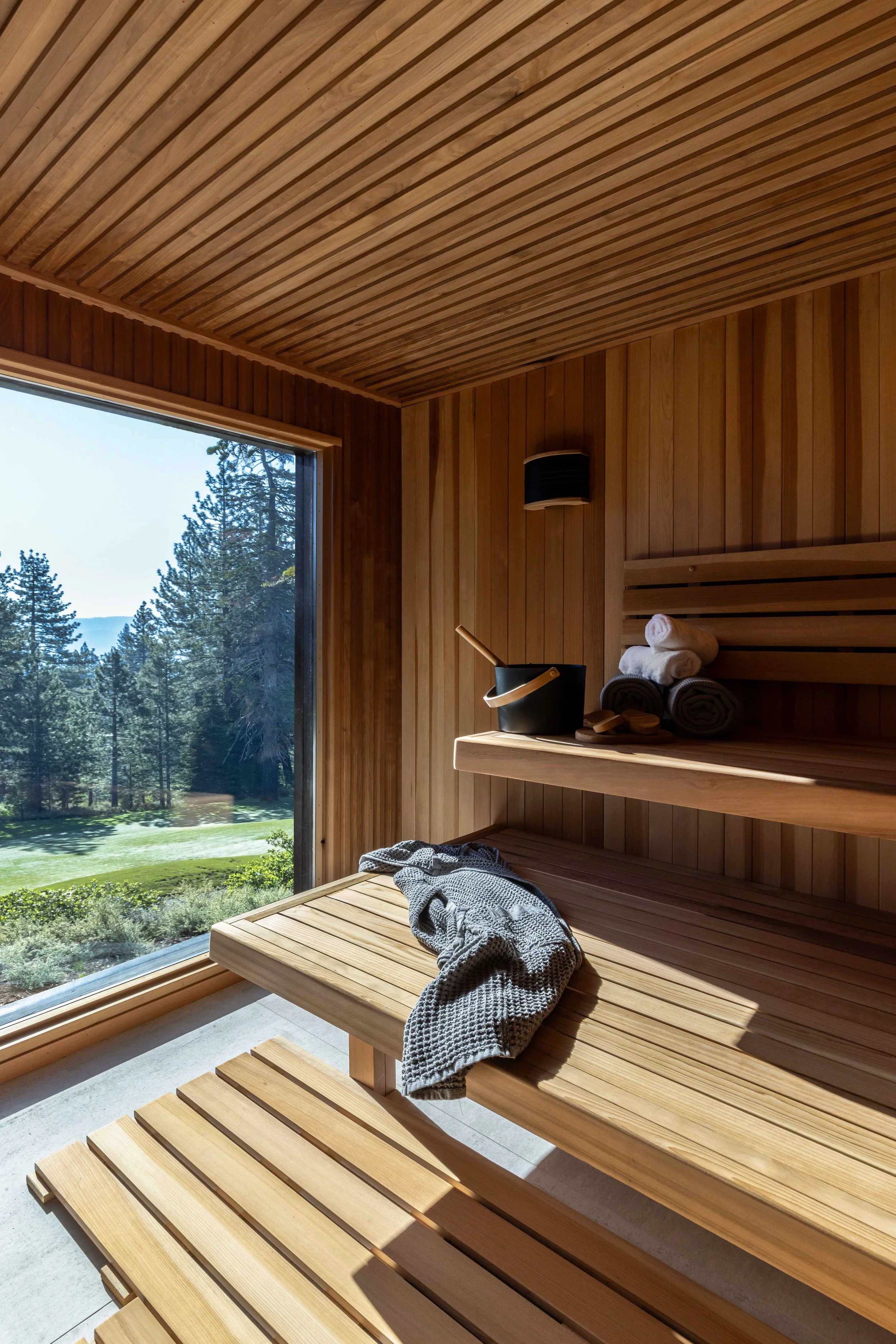 A wooden sauna room with a bench and a roll of towels, a sauna bucket, and a ladle, with a large window looking out on a forested landscape.