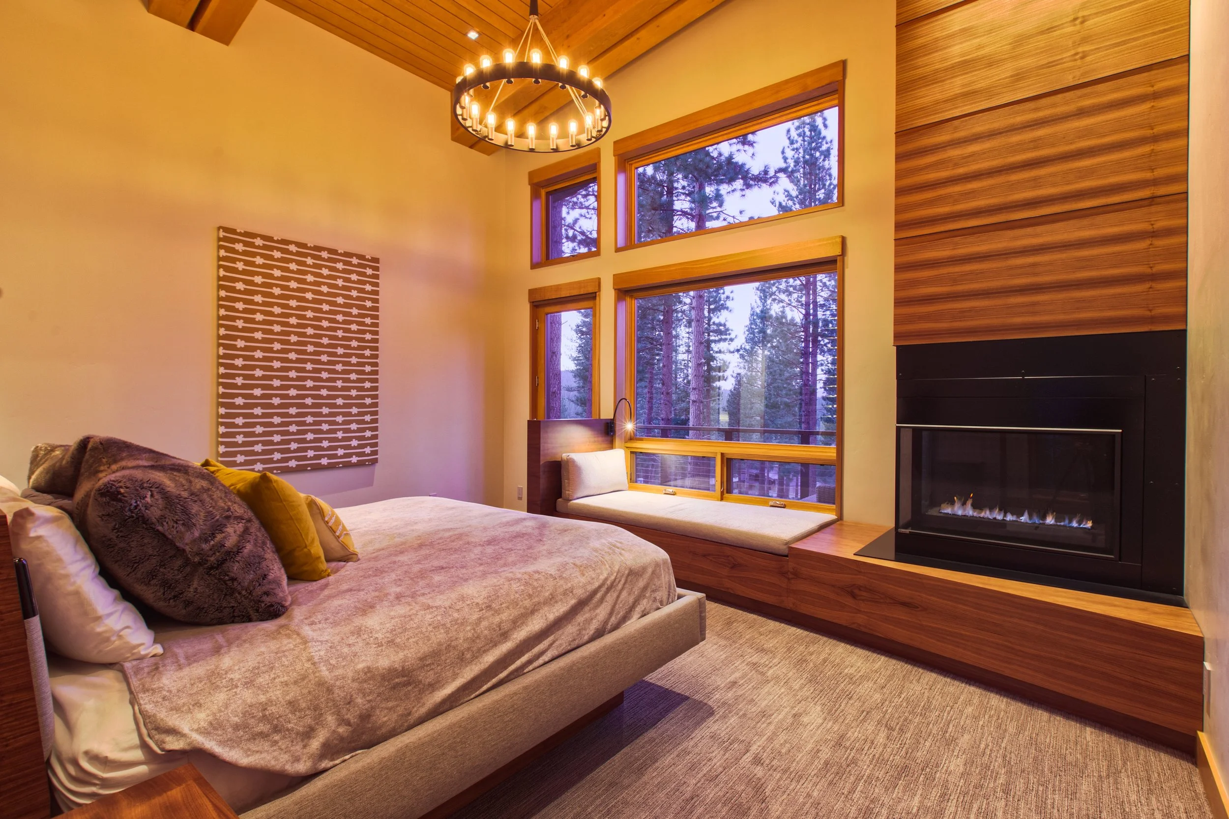 Interior of a bedroom with large windows showing a forest view, a bed with multiple pillows, a built-in window seat, a modern fireplace, and a chandelier hanging from a wooden ceiling.